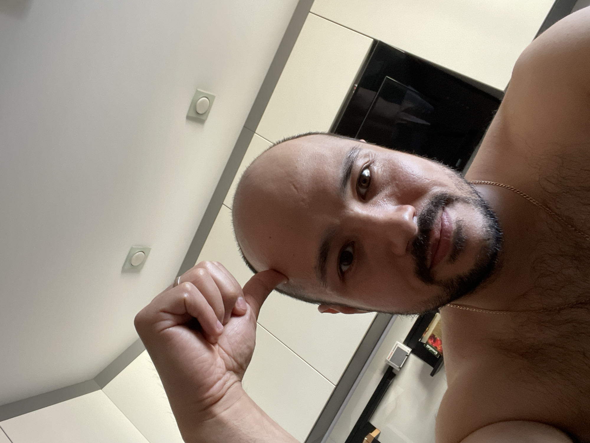 A close-up, low-angle selfie photograph shows a balding man with a dark beard touching his temple with his index finger. He is shirtless and wearing a gold chain. The background includes a white ceiling with square recessed lights, white kitchen cabinets, and a black built-in oven. The location is not discernible from the image and cannot be determined to be in Izhevsk, Russia.