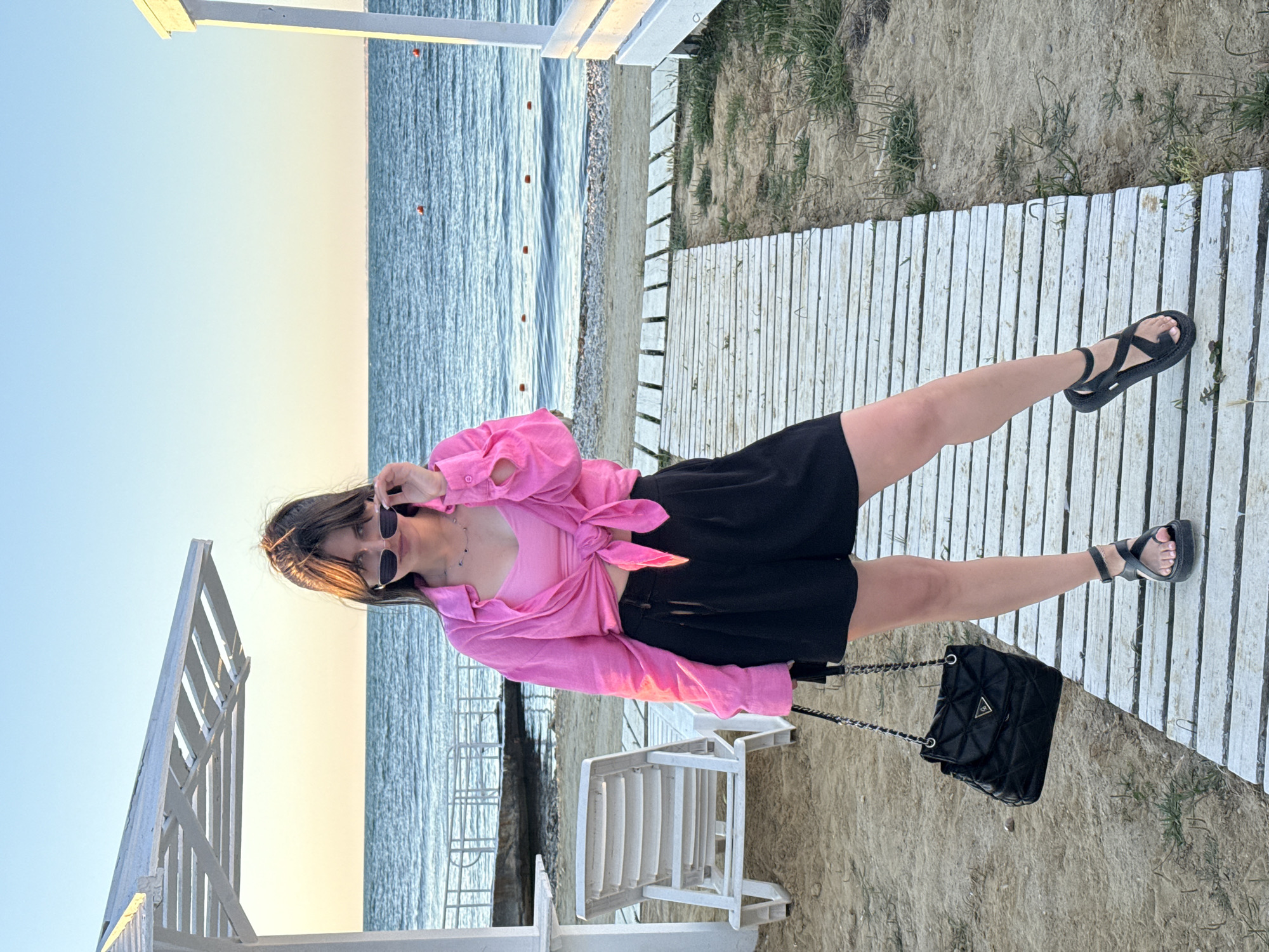 A young woman stands on a wooden boardwalk at the beach, facing forward and looking towards the camera. She is wearing a bright pink knotted shirt over a pink top, black shorts, and black sandals. She is also holding a black quilted handbag. The woman is wearing sunglasses and has her hair pulled up. The boardwalk leads towards the ocean, which is visible in the background. There are lounge chairs and white structures on either side of the boardwalk. The sky is clear and blue.