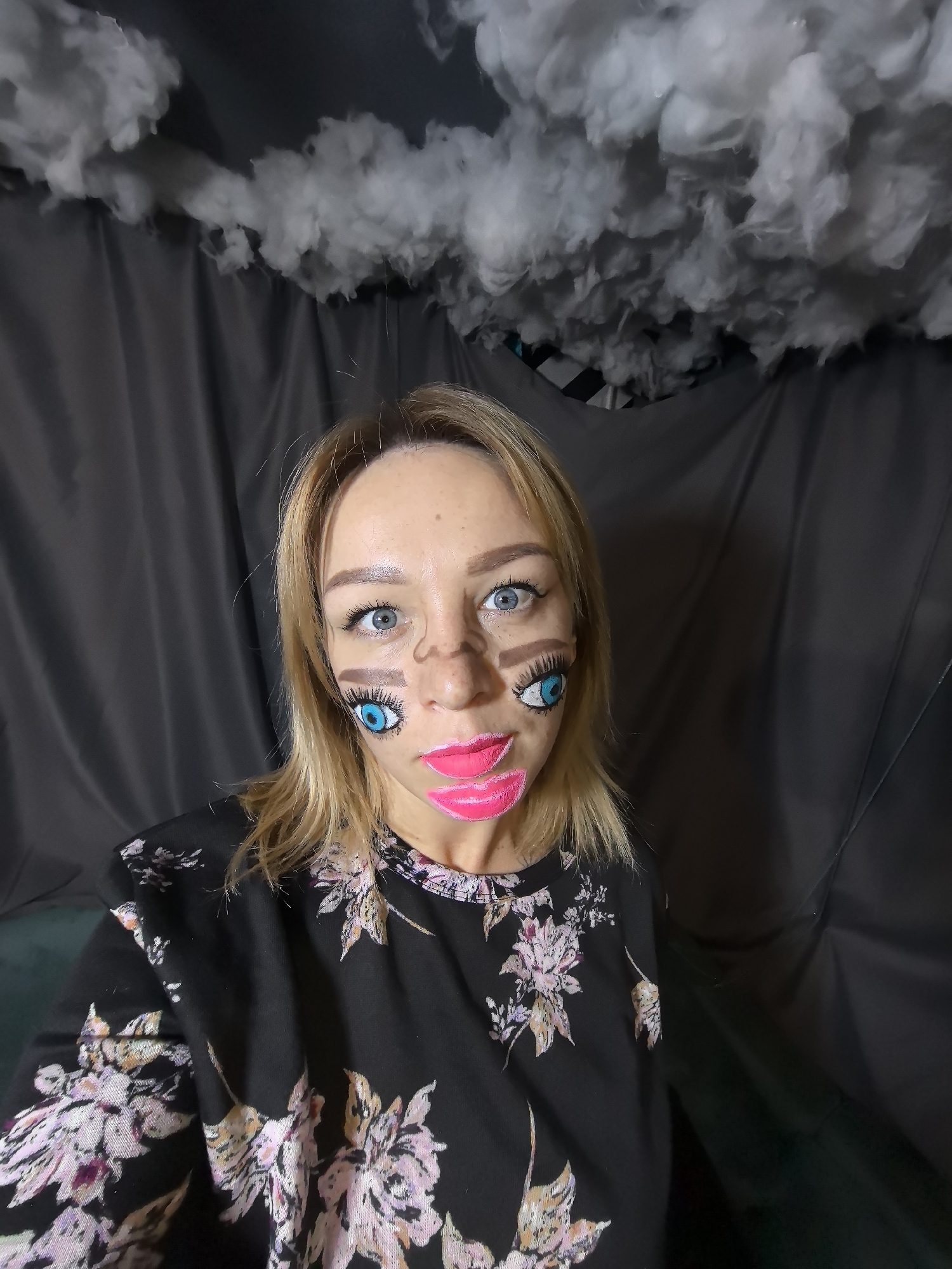 A medium close-up, eye-level shot captures a woman indoors, possibly in Noviy, Russia. The woman, appearing to be in her 20s or 30s, has blonde hair and striking light blue eyes that are wide open and looking directly at the camera. She is adorned with elaborate, doll-like face paint. This makeup includes large, bright blue eyes with white highlights and black outlines painted onto her cheeks, beneath her natural eyes. Thick, rectangular brown eyebrows are painted below her natural brow line, and her lips are covered in vibrant, glossy pink paint that extends beyond her natural mouth onto her chin. A light brown, drawn line also crosses the bridge of her nose.
She is wearing a black long-sleeved top with a floral pattern featuring various shades of pink, purple, white, and orange flowers and leaves.
The background consists of dark gray or black draped fabric, creating a simple, enclosed setting. Along the top of the frame, several textured white cloud-like decorations, likely made of cotton or a similar soft material, are suspended against the dark backdrop. A small hint of a dark green or teal surface is visible at the very bottom left corner. The lighting appears to be artificial, typical of an indoor setting. The elaborate face paint suggests she is participating in a costume event, a themed photoshoot, or creating artistic content. No text is visible in the image.