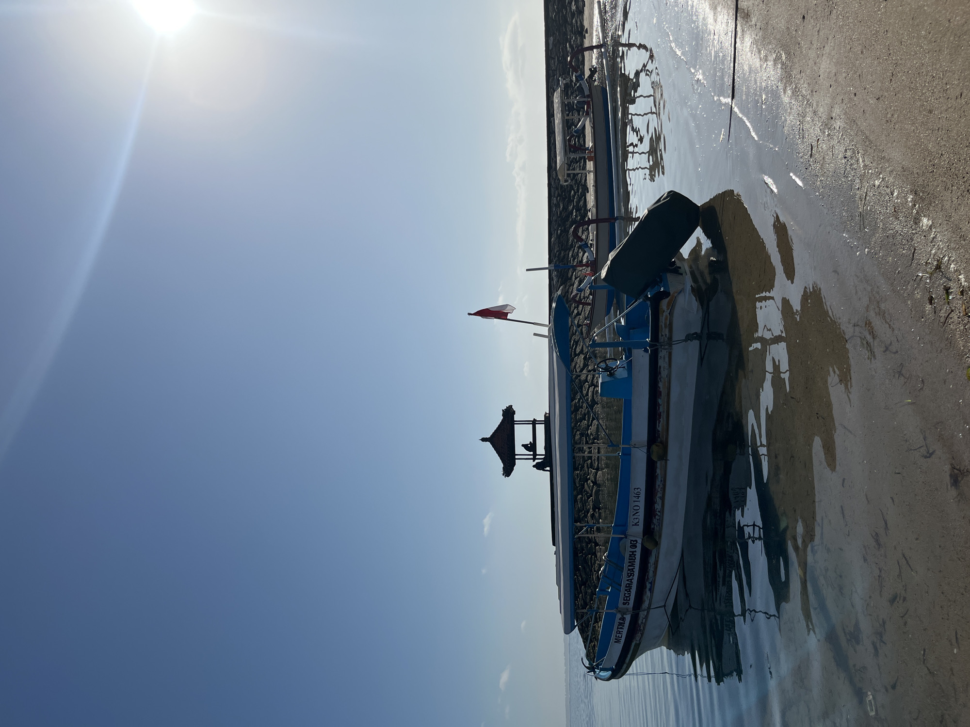 In Kuta Selatan, Indonesia, this image captures a serene beach scene bathed in bright sunlight. Two traditional Jukung boats are partially submerged in calm, shallow waters near the sandy shore. The boat in the foreground, painted white with blue accents, displays text that reads "MERRIKA SEGARA SAMUH 03 K3 NO 1463." A dark green tarp is draped over a portion of this boat. Behind it, another Jukung boat, this one with a white canopy, is moored. A red and white Indonesian flag flutters from a pole on the second boat. In the background, a small, dark-roofed gazebo sits atop a rocky breakwater, with two silhouetted figures visible inside. The sky is a clear, brilliant blue with a bright sun casting a lens flare in the upper right corner.