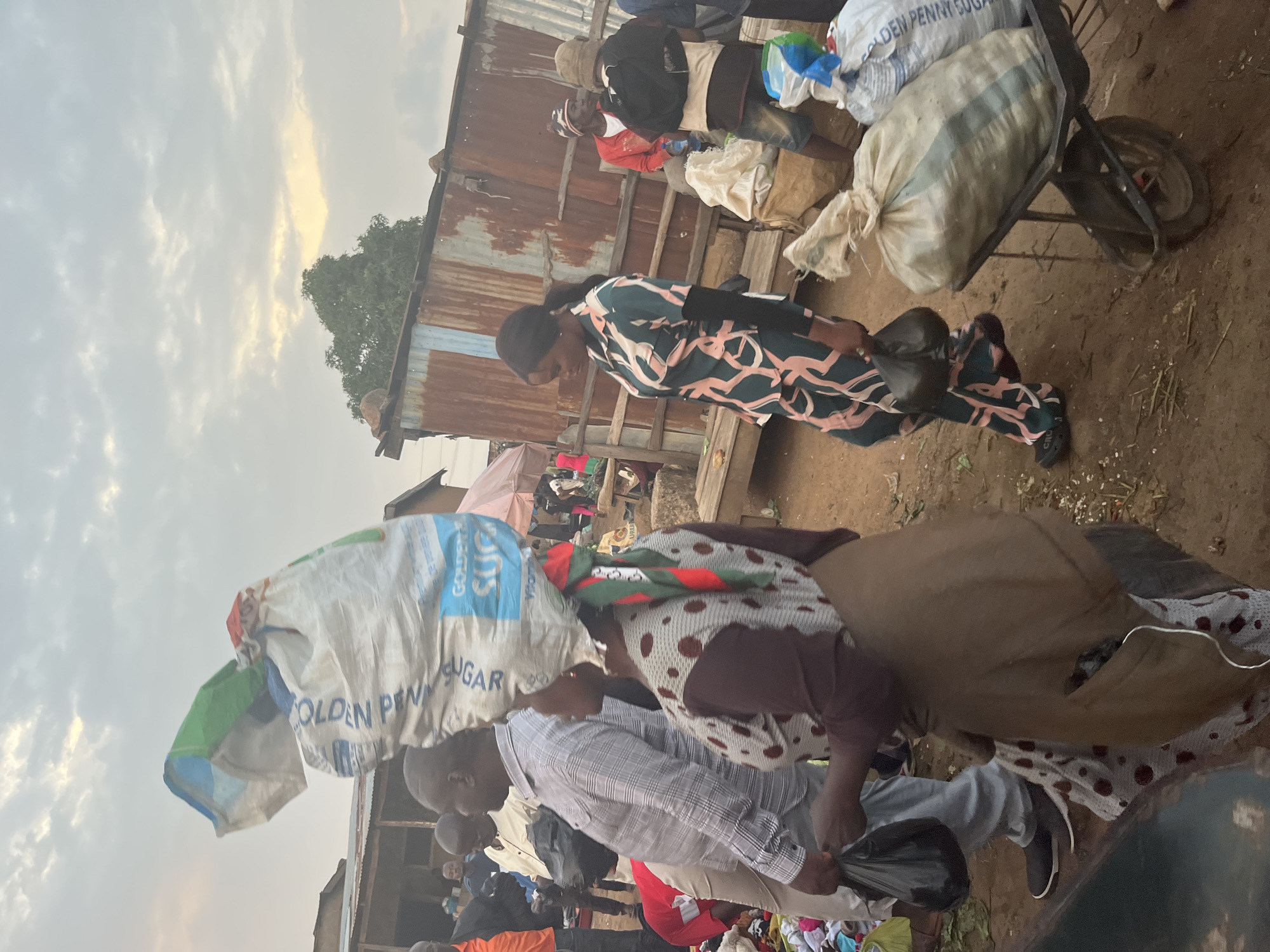 The image depicts a bustling outdoor market scene in Gura Top, Nigeria, under a cloudy sky. In the foreground, a woman walks with a large bag of what appears to be sugar balanced on her head. The bag is made of a white, woven material and has green and blue plastic attached to its opening. She is wearing a brown headscarf with red and green accents, and a brown and white patterned dress. Beside her, a man in a grey plaid shirt and light-colored pants also carries a black plastic bag.
Further into the market, a woman in a green and pink patterned outfit walks with a black plastic bag in her hand, near a wheelbarrow filled with several large sacks. Behind her, two men are visible; one in a red jacket and beanie, and another in a hat and backpack. The background reveals stalls constructed from corrugated metal and wood, with various goods and people milling about. The ground is dusty and appears to be unpaved, with some scattered debris. The overall impression is one of a lively and active marketplace.