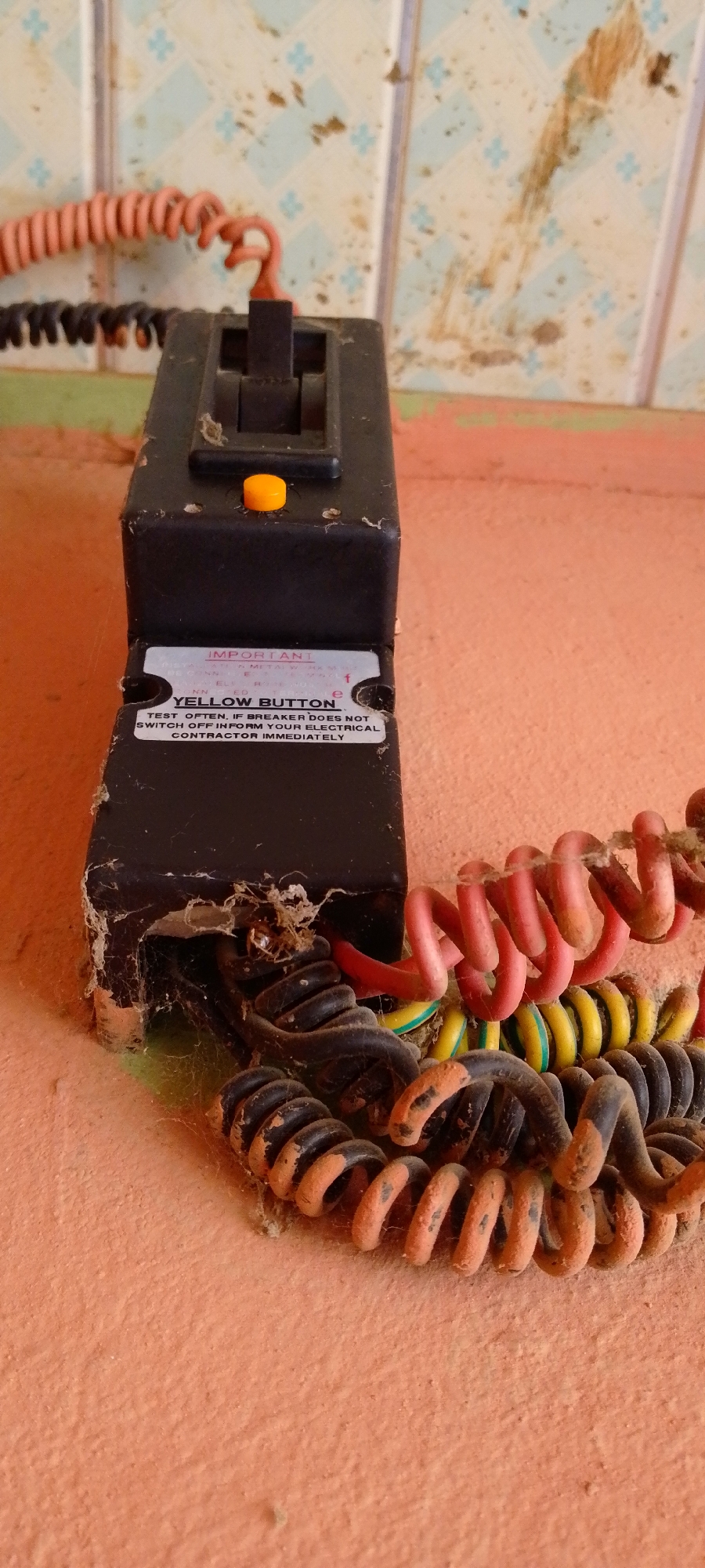 The image shows a close-up, eye-level view of an old, black electrical breaker switch with a yellow test button, mounted on an orange surface that appears to be a wall. The breaker is dusty and has some cobwebs around it, especially where the coiled electrical wires enter the device. The wires are black, red, and yellow. The wall behind the breaker has a light-colored tiled pattern with blue accents and is stained with brown marks. The scene suggests an indoor location, possibly a residential or commercial building, and given the request's context, it is assumed to be in Jalingo, Nigeria. There are no people visible in the image.