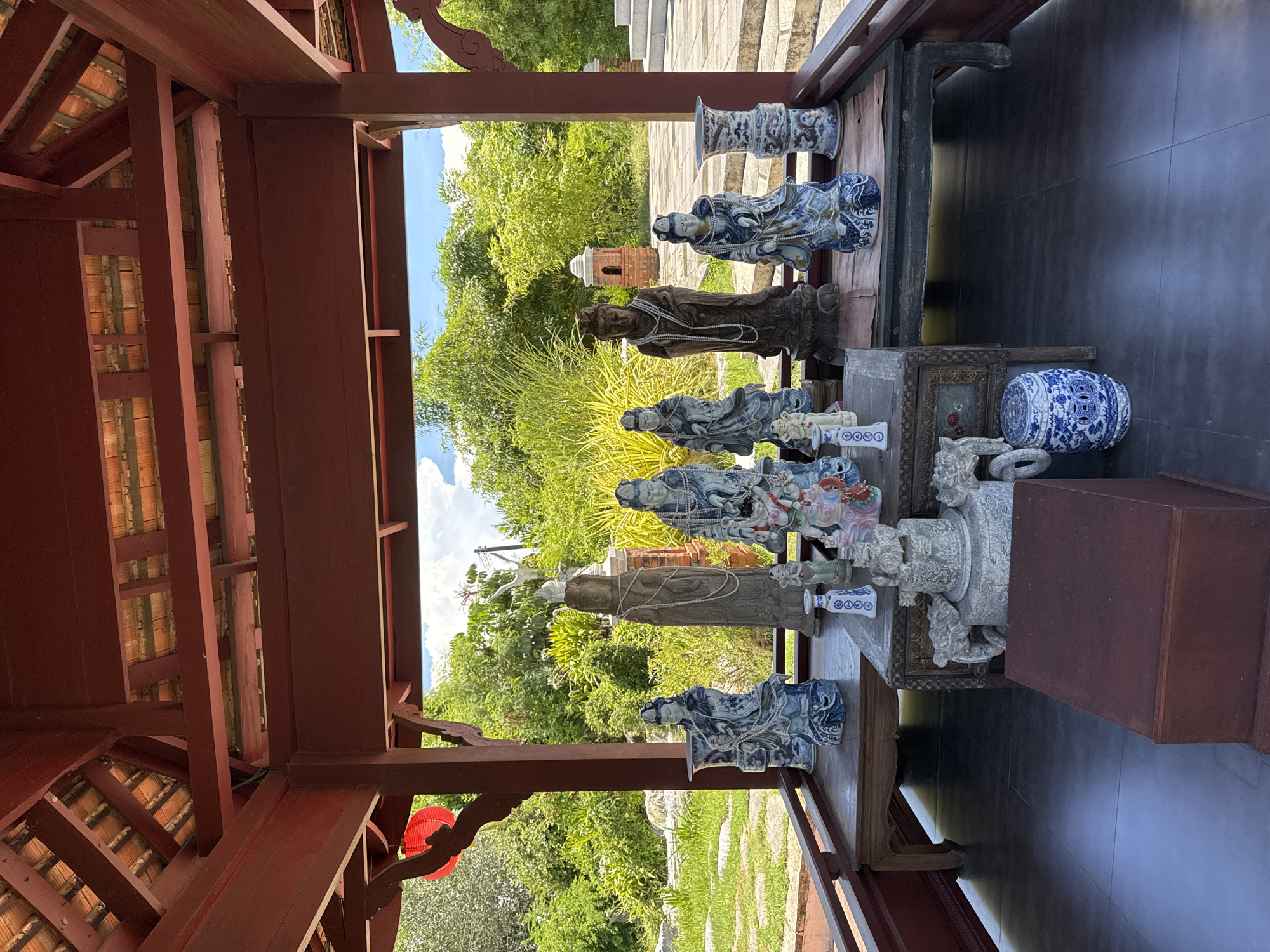 The image depicts a collection of statues and vases displayed on a wooden table under a red wooden structure, possibly a pavilion. The statues are predominantly blue and white porcelain, depicting reclining figures, while the vases are of various shapes and patterns. A brown, standing statue is also present. The background shows a lush green outdoor scene with trees and plants, suggesting a garden or park setting. The location is in the city of Bang Pu Subdistrict Municipality, Thailand. There are no people visible in the scene.