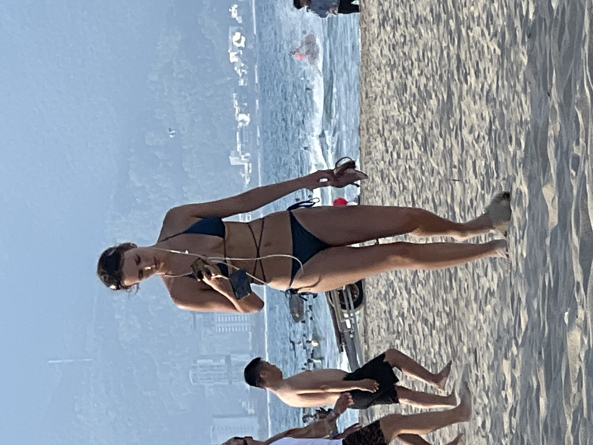 A woman in a dark blue bikini walks on a sandy beach, looking down at her phone. She is holding a pair of sandals in her right hand. Behind her, the ocean is visible with several people swimming and a jet ski speeding across the water. In the distance, a city skyline with tall buildings and mountains can be seen under a hazy sky. The scene appears to be located in Đà Nẵng, Vietnam, a coastal city known for its beaches and urban landscape.