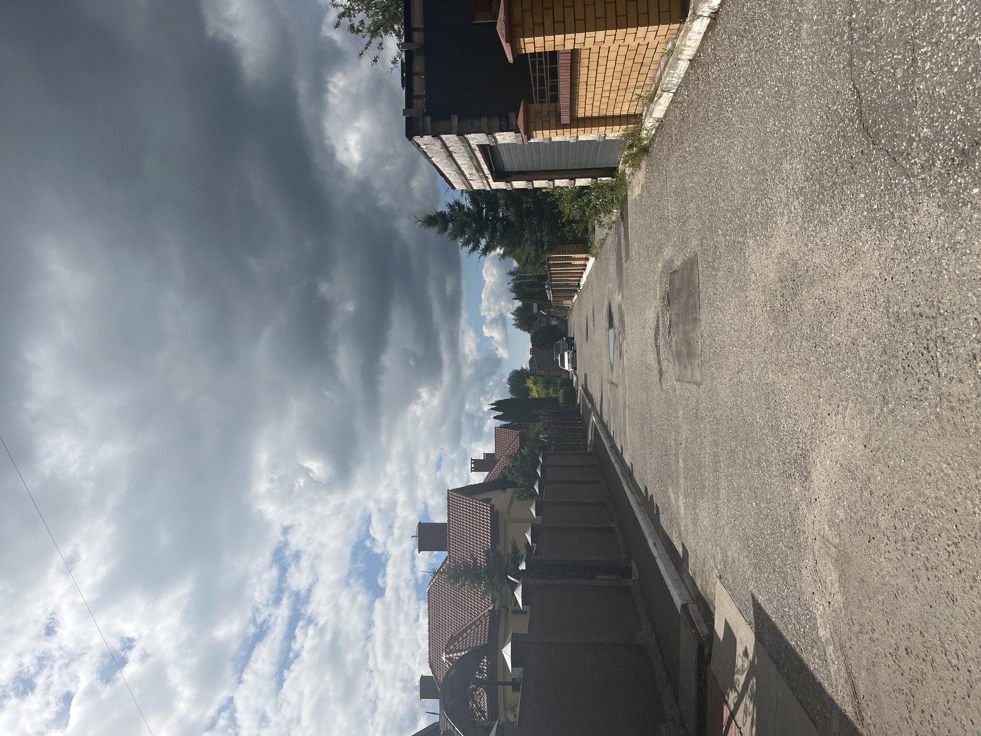 The image shows a residential street in the city of Orekhovka, Russia. The street is paved with asphalt and lined with houses and fences. On the left side of the street, there are several houses with red tiled roofs and brick fences. On the right side, there is a garage with a dark gray door, a yellow brick wall, and a fence. The street is empty except for a white car parked in the distance. The sky is partly cloudy with dark gray storm clouds. There are trees and greenery visible along the street.