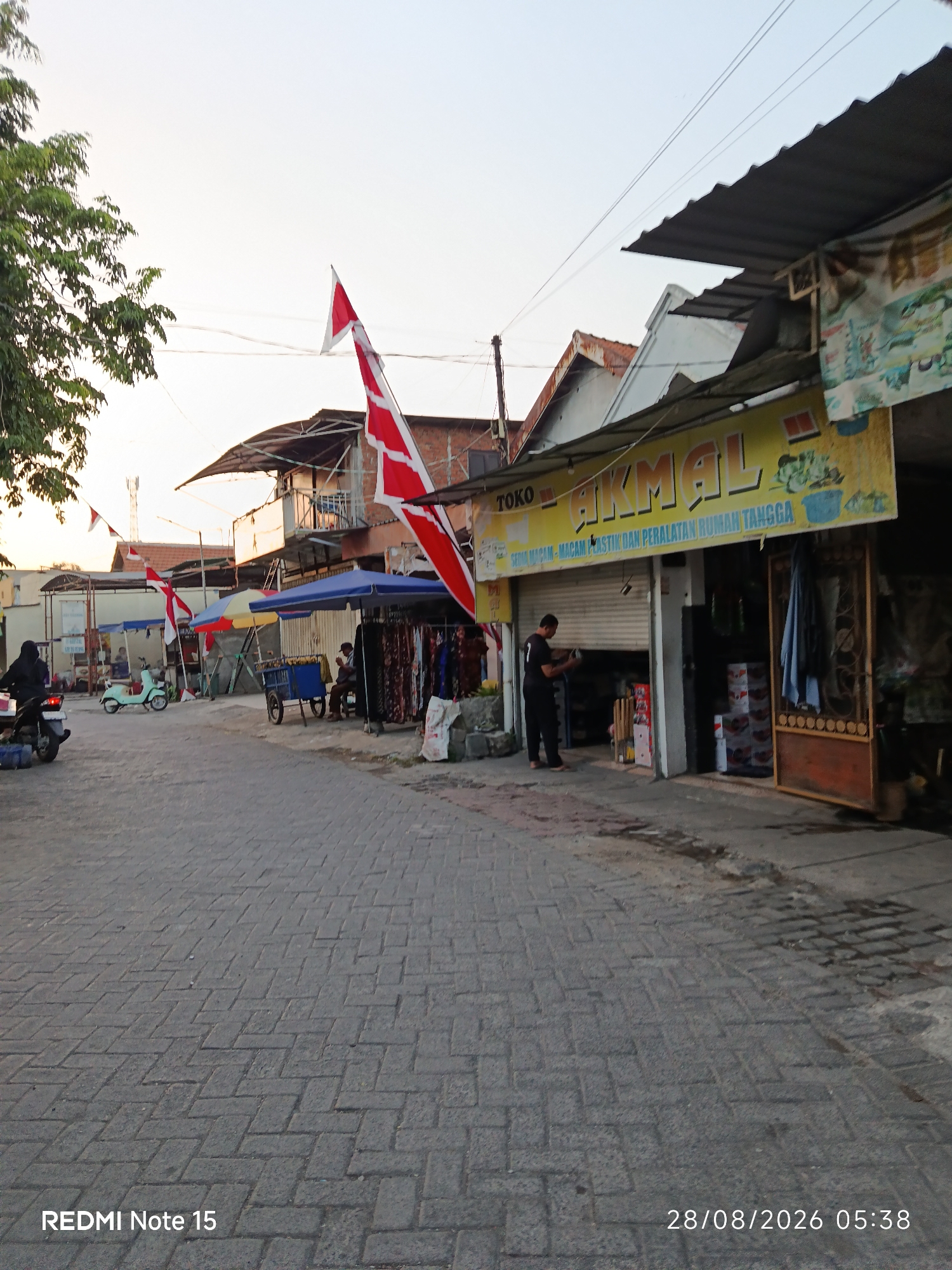 In the city of Keboansikep, Indonesia, a street scene unfolds at dawn. The road is paved with interlocking gray bricks, arranged in a herringbone pattern. On the right side of the street, a shop with a yellow awning displays the text "TOKO AKMAL" in large, bold, red and white letters, with smaller text beneath reading "SEDIA MACAM-MACAM PLASTIK DAN PERALATAN RUMAH TANGGA" (selling various plastic and household items). The shop has a metal rolling door that is partially open, revealing shelves of merchandise inside. A man in a black t-shirt and black pants stands in the doorway, facing the interior. Adjacent to the shop, a rustic wooden door with intricate carvings stands open, revealing a glimpse of the interior.
Further down the street, on the left, a woman wearing a black hijab and black clothing rides a motorcycle. To her right, a light green scooter is parked. Several market stalls with blue and yellow umbrellas are set up along the side of the road, displaying various goods, including clothing on racks and a cart filled with produce. Two men are seated near the stalls, one appearing to interact with the merchandise. Red and white Indonesian flags are prominently displayed on poles, adding a patriotic touch to the scene. In the background, a cellular tower is visible against the pale sky. The overall atmosphere suggests a bustling market area beginning its day.