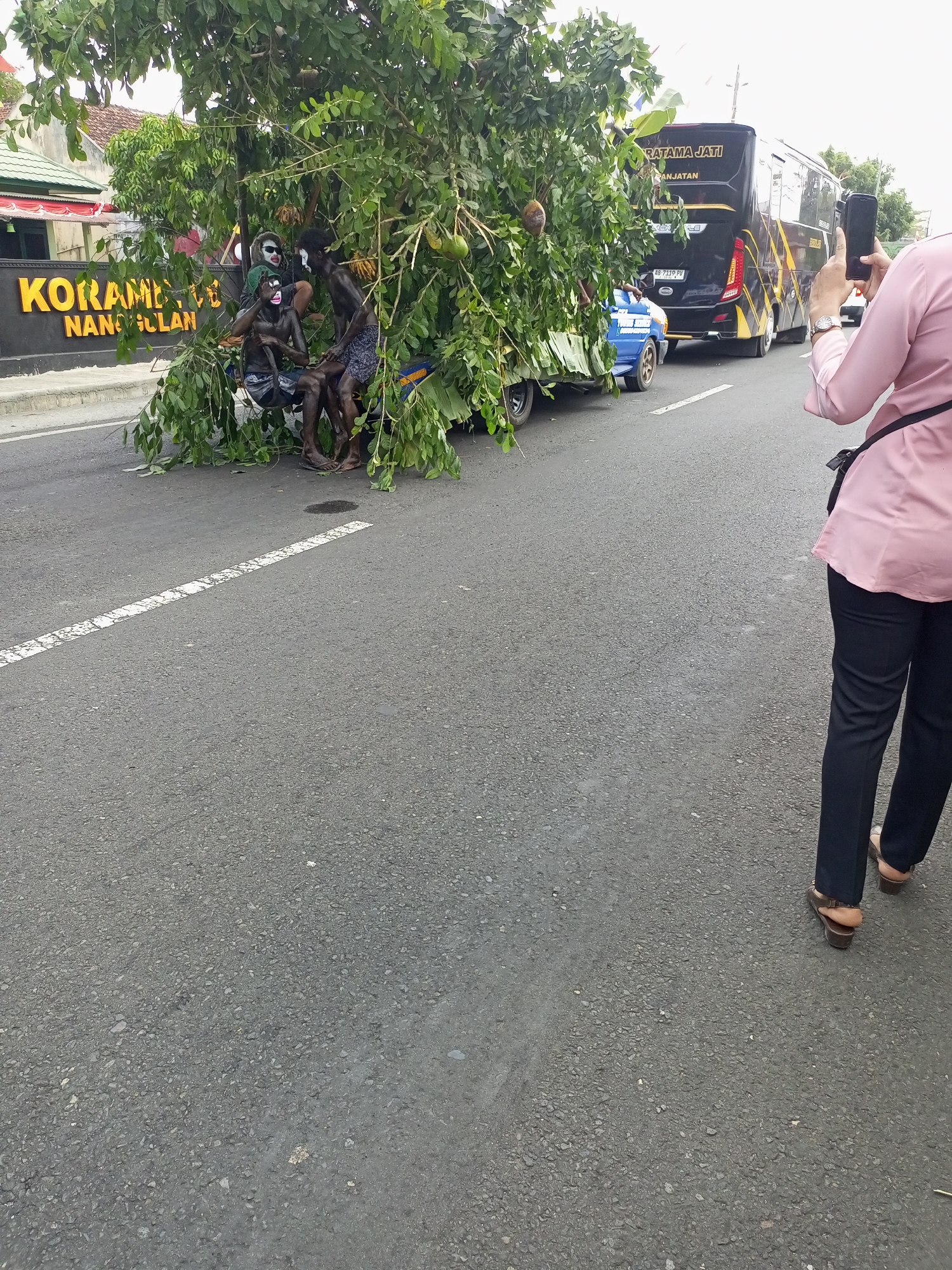 The image depicts a scene on a road in Nanggulan, Indonesia. In the foreground, a person wearing a pink shirt and black pants is holding a smartphone and taking a picture. To the left, a vehicle is heavily laden with branches and foliage, obscuring its contents. Within the foliage, three individuals are visible, appearing to be covered in dark paint or makeup, with white markings on their faces. One individual wears glasses and has dark shorts, another has patterned shorts, and a third has plain shorts. Behind this vehicle, a large black bus with yellow accents and the text "PRATAMA JATI" is stopped on the road. A small blue vehicle is positioned between the bus and the foliage-laden vehicle. The environment appears to be an urban or semi-urban setting with buildings and signage visible in the background. The road surface is asphalt, marked with white lines.