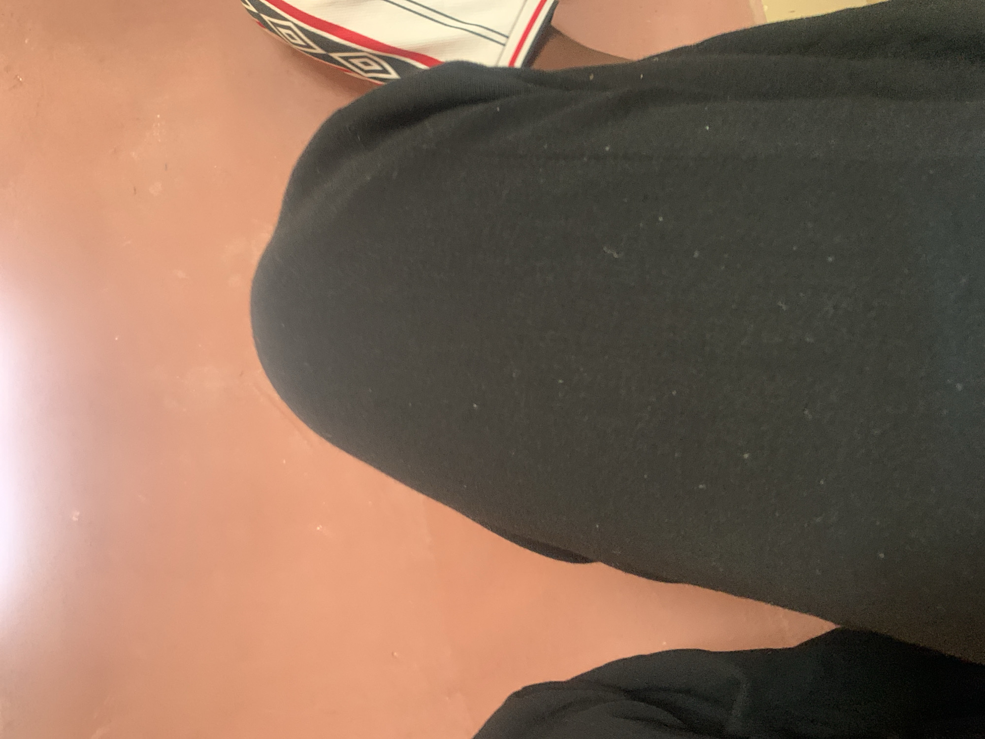 The image shows a close-up view of two individuals from the waist down, seated in a room. The wall behind them is a peachy-pink color, with some visible texture and light dust specks.
The individual on the left is wearing black clothing. The individual in the center is also wearing black clothing that covers their legs and is draped over their lap.
The individual on the right is wearing a white shirt with a black and red geometric pattern on the sleeve. The pattern consists of diamond shapes outlined in black and filled with red and black.
The scene appears to be indoors, likely in a residential setting in Jauro Yinu, Nigeria, given the context. The lighting suggests an indoor environment, possibly during the daytime. There are no overt indications of any particular activity beyond sitting.