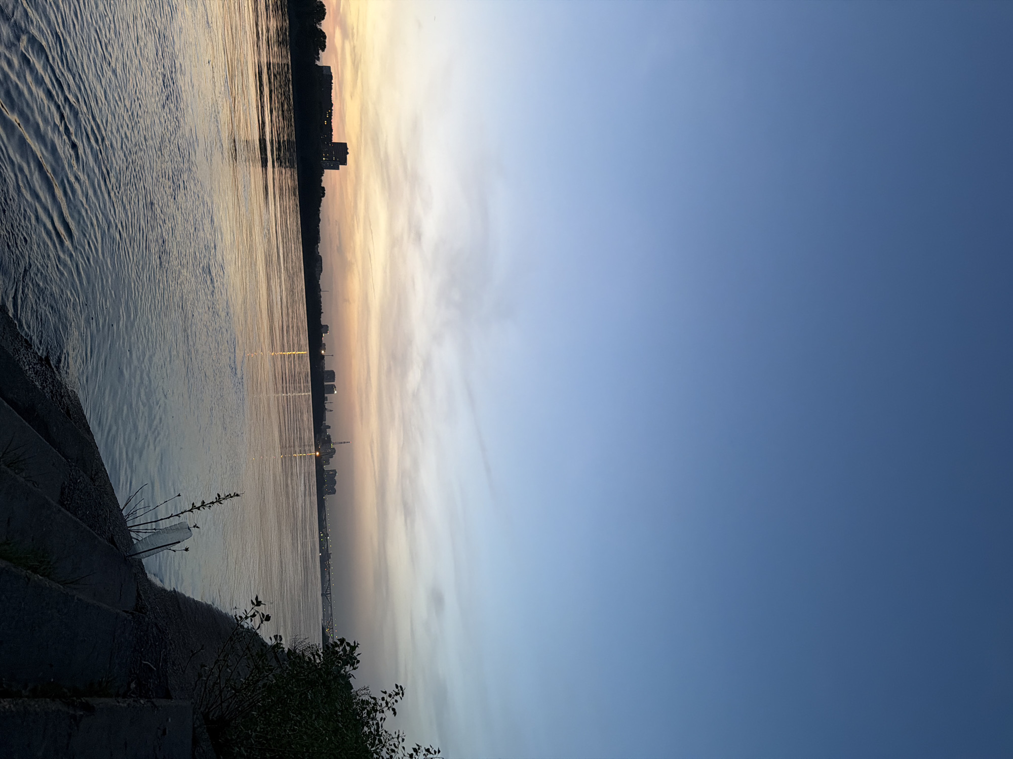 The image shows a view of the Oka River in Nizhny Novgorod, Russia, at sunset or sunrise. The foreground consists of concrete steps leading down to the riverbank, with some vegetation growing on the steps. The river reflects the colors of the sky, primarily gold and orange hues near the horizon. Across the river, the skyline of Nizhny Novgorod is visible, including buildings and a bridge lit up in the distance. The sky transitions from the warm colors of the horizon to a clear blue. There are no people visible in the image.