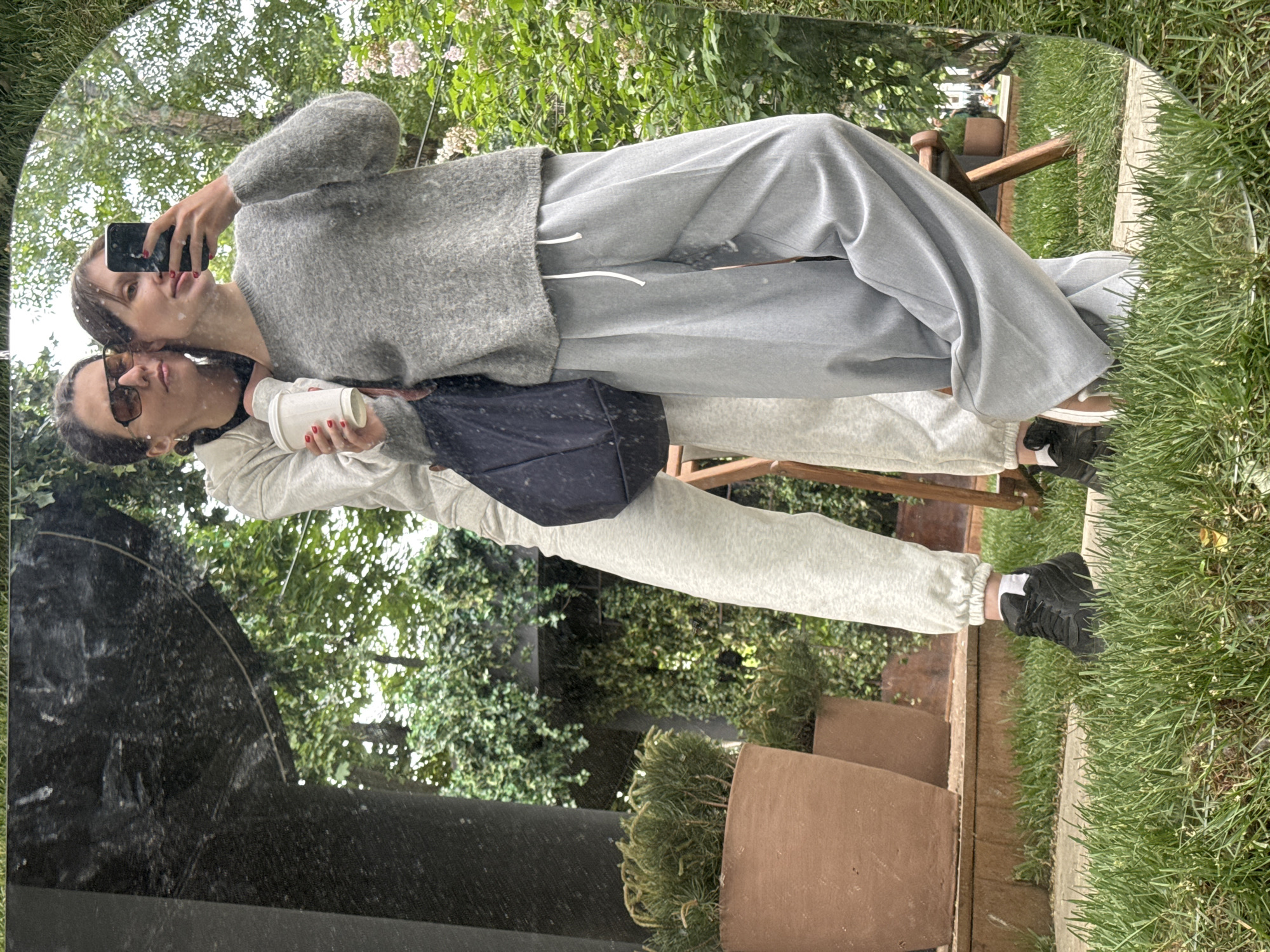 The image, displayed rotated 90 degrees clockwise, captures a mirror selfie of two women in an outdoor setting.
The two main subjects are women reflected in a large, slightly curved mirror. The woman positioned towards the upper part of the image is taking the selfie, holding a black smartphone with her right hand. She has dark hair, wears a gray knit sweater and light gray, wide-leg sweatpants, and has red nail polish. Her head is tilted slightly. The second woman stands just behind and to the right of the first, wearing sunglasses pushed up on her head or just above her eyes, and holding a white cup in her right hand. She is dressed in a light gray hooded top and matching light gray sweatpants. A dark-colored shoulder bag is slung across her body. Both women wear dark sneakers with white socks.
The activity taking place is a casual mirror selfie, likely to capture a moment outdoors.
The setting appears to be a lush garden or patio, consistent with the Moscow, Russia, location context. The background, visible in the mirror's reflection, is dominated by vibrant green foliage, including trees, bushes, and grass. Some small white or light pink flowers are scattered among the greenery. Terracotta-colored potted plants are visible at the bottom of the mirror, and parts of a wooden chair or outdoor furniture can be seen in the reflection. The lighting is natural and bright, suggesting daytime, possibly morning or afternoon, with no harsh shadows indicating an overcast or diffused sky. The women appear relaxed and comfortable. The mirror surface shows some smudges. There is no visible text in the image.