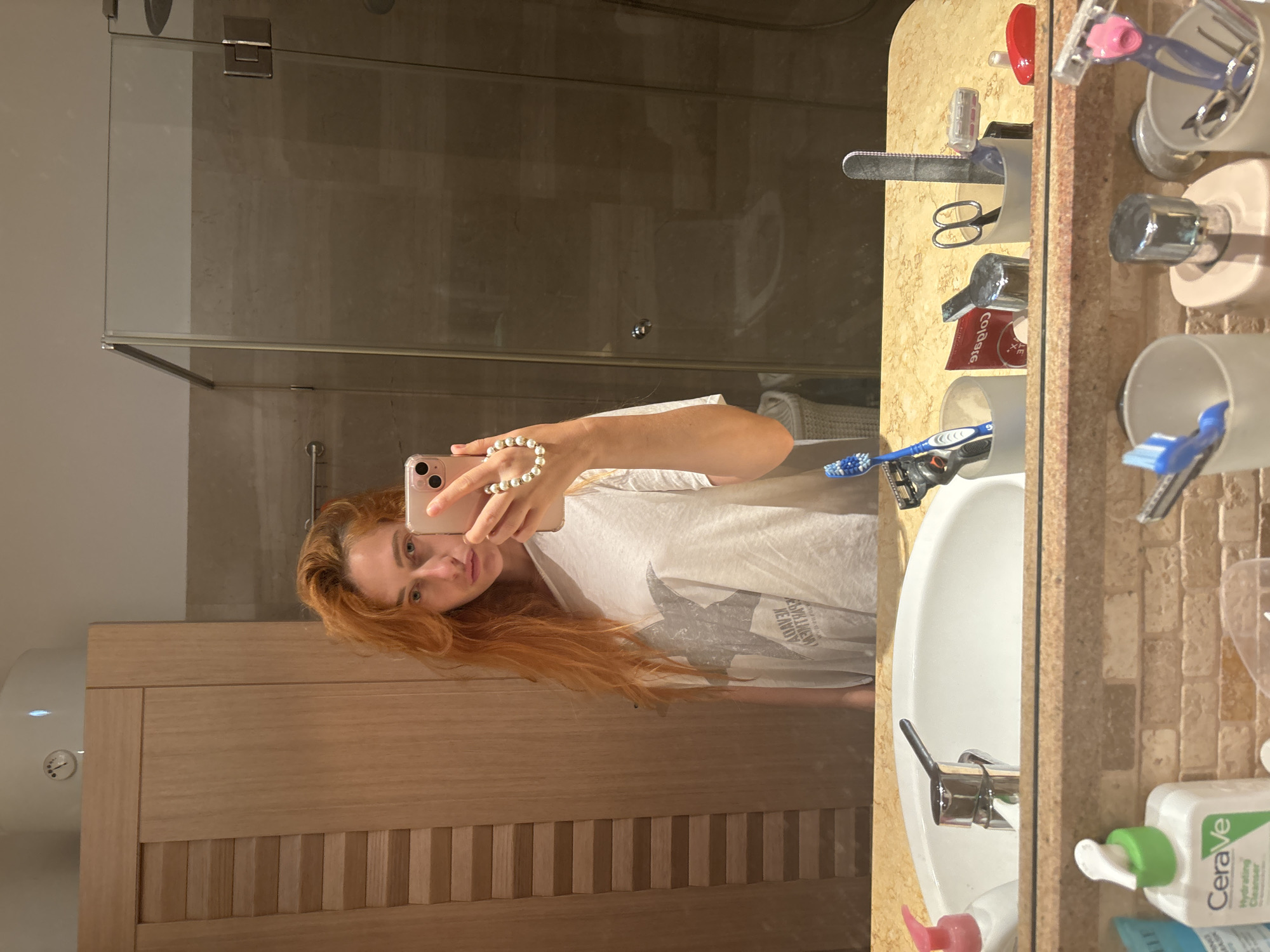 In Budva, Montenegro, a young woman with long, red hair stands in a well-lit bathroom, taking a mirror selfie. She is wearing an oversized white t-shirt with a grey star graphic. Her right hand, adorned with a pearl bracelet, holds up a pink smartphone, capturing her reflection. The bathroom features a wooden cabinet to the left, a modern sink with a chrome faucet, and a glass-enclosed shower to the right. The countertop is made of a speckled beige material, organized with various toiletries, including toothbrushes, razors, and a CeraVe cleanser.