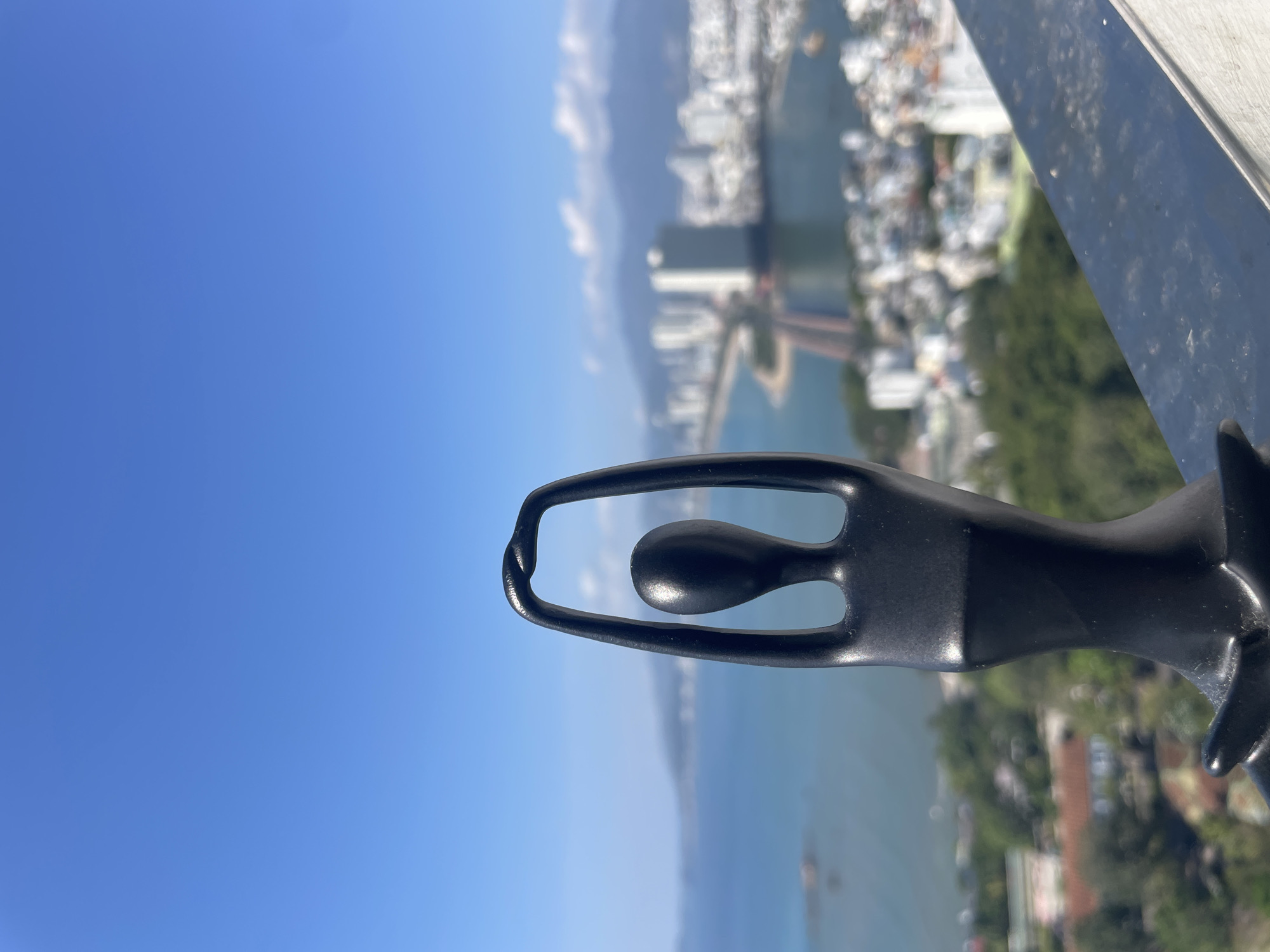 A high-angle, medium shot presents a black, stylized metal figure of a person with arms raised, positioned against a blurred background of the Nha Trang cityscape in Bac Nha Trang Ward, Vietnam. The background shows a coastal city with buildings, a body of water, and a clear blue sky. The figure appears to be part of a railing or decorative element on a balcony or high vantage point.