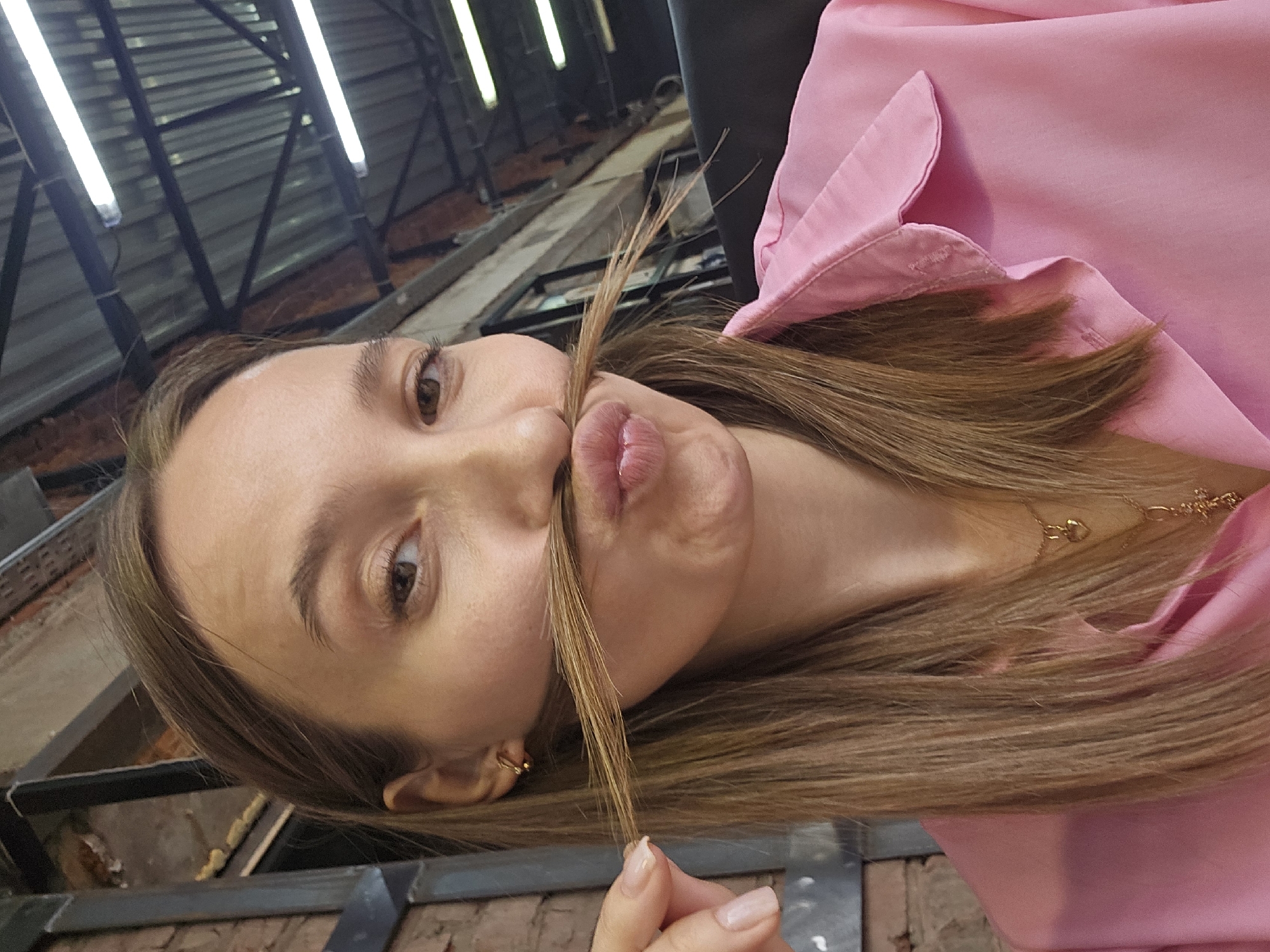 A young woman with light brown hair and light skin, wearing a pink shirt, is holding a strand of her hair up to her upper lip, creating a makeshift mustache, while making a pursed-lip expression. She is indoors, and in the background, there is a metal structure, possibly a ceiling or part of a building's framework, with fluorescent lighting. The environment appears to be a public space or building undergoing renovation or construction, potentially in Chaikovsky, Russia, though there is no overt visible signage or landmarks confirming the exact location.