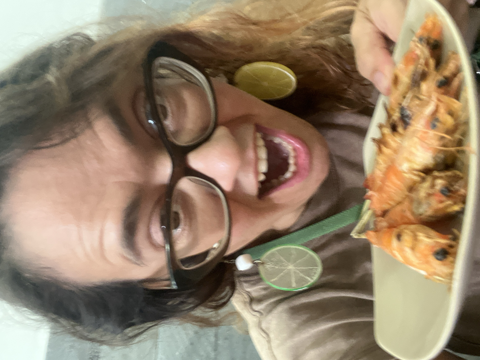 A close-up, selfie-style photo shows a person with medium-length, wavy, light brown hair and glasses with thick, dark frames. The person is wearing a brown t-shirt and has large, circular earrings that appear to be slices of lime. Their mouth is open, revealing teeth and pink lips. In the foreground, a white plate holds several grilled shrimp. The background is blurred and appears to be an indoor setting, possibly a restaurant or home. The photo is taken at a slightly downward angle, with the plate of shrimp being the closest object to the camera.