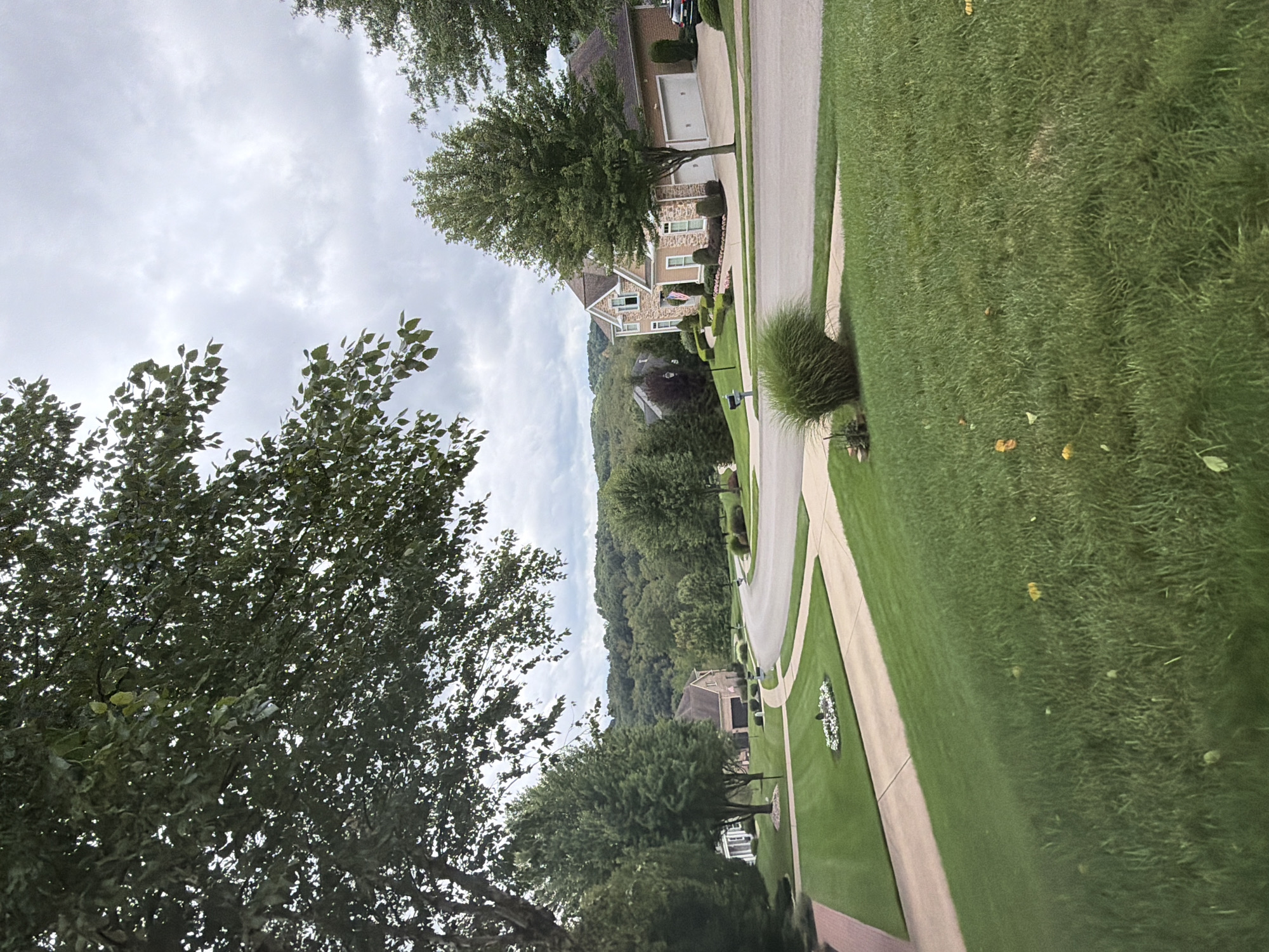 The image depicts a suburban street scene in Wexford, United States of America, under an overcast sky. Lush green lawns, meticulously manicured, dominate the foreground and the sides of the paved road that curves through the neighborhood. A large tree with dense foliage partially obscures the upper left portion of the frame.
Several large, well-maintained houses are visible, featuring architectural details such as stone accents, multiple windows, and attached garages. One prominent house, with a beige and stone facade, is situated on the right side of the road. It has a steeply pitched roof and is surrounded by neatly trimmed bushes and landscaping. An American flag is displayed on its porch.
The road itself is a light gray asphalt, with clearly defined lanes and curbs. Sidewalks are present along some sections of the street, also bordered by grass. A mailbox stands near the edge of one of the lawns. In the background, rolling hills covered in dense trees create a picturesque backdrop. The overall impression is one of affluence and a well-kept residential community. There are no people visible in the image.