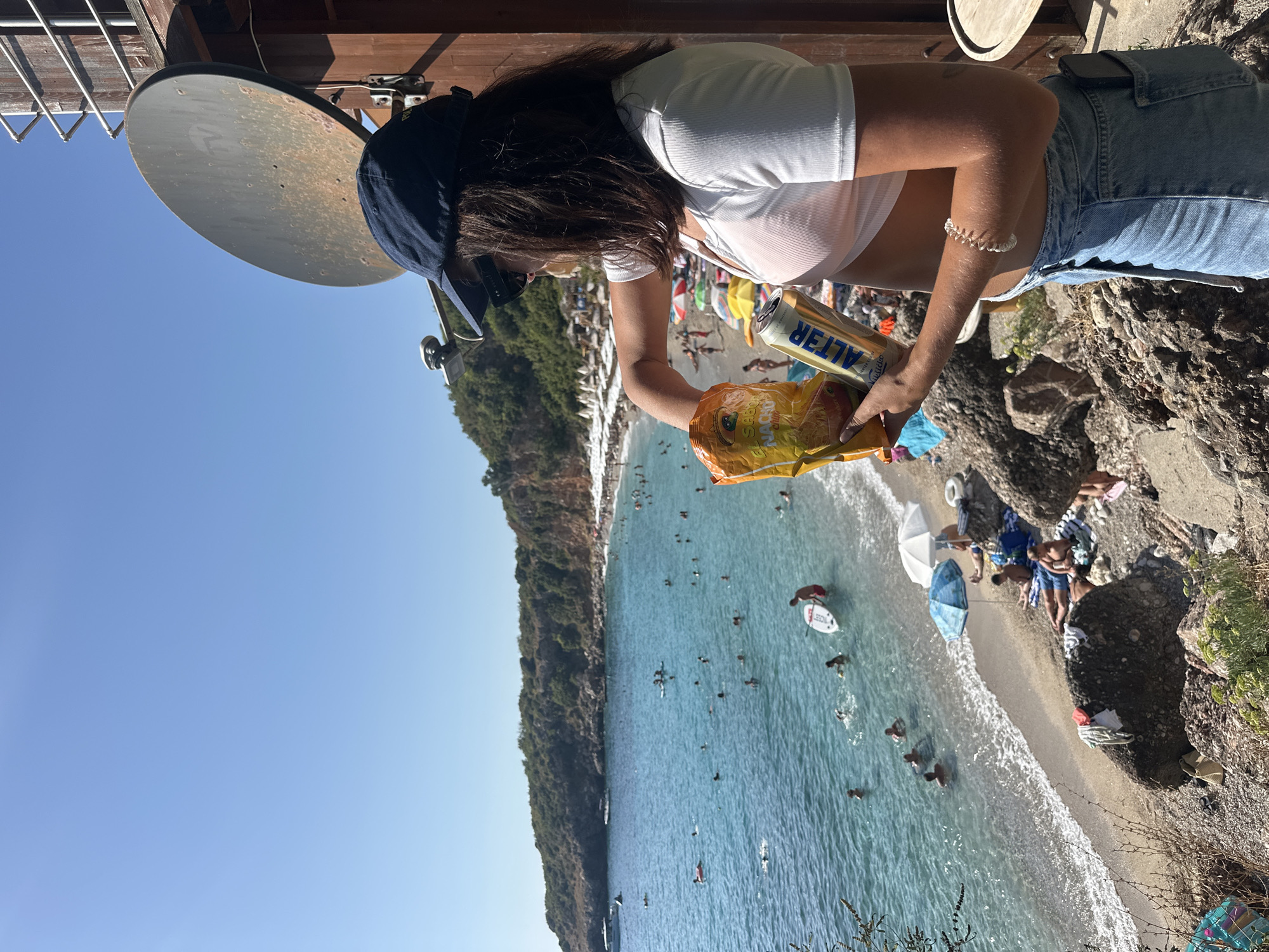 An exterior, medium shot captures a young woman wearing a navy blue baseball cap, sunglasses, and a white crop top over an orange bikini top, holding a bag of chips and a beer can. She is standing on a rocky, elevated viewpoint overlooking a crowded beach and a body of clear turquoise water. The beach is lined with white umbrellas and beach chairs, and numerous people are swimming, sunbathing, and paddling on boards in the water. Green hills form the backdrop, and a satellite dish is mounted on a wooden structure to the woman's right. The scene is set in Drobnići, Montenegro, on a sunny day.