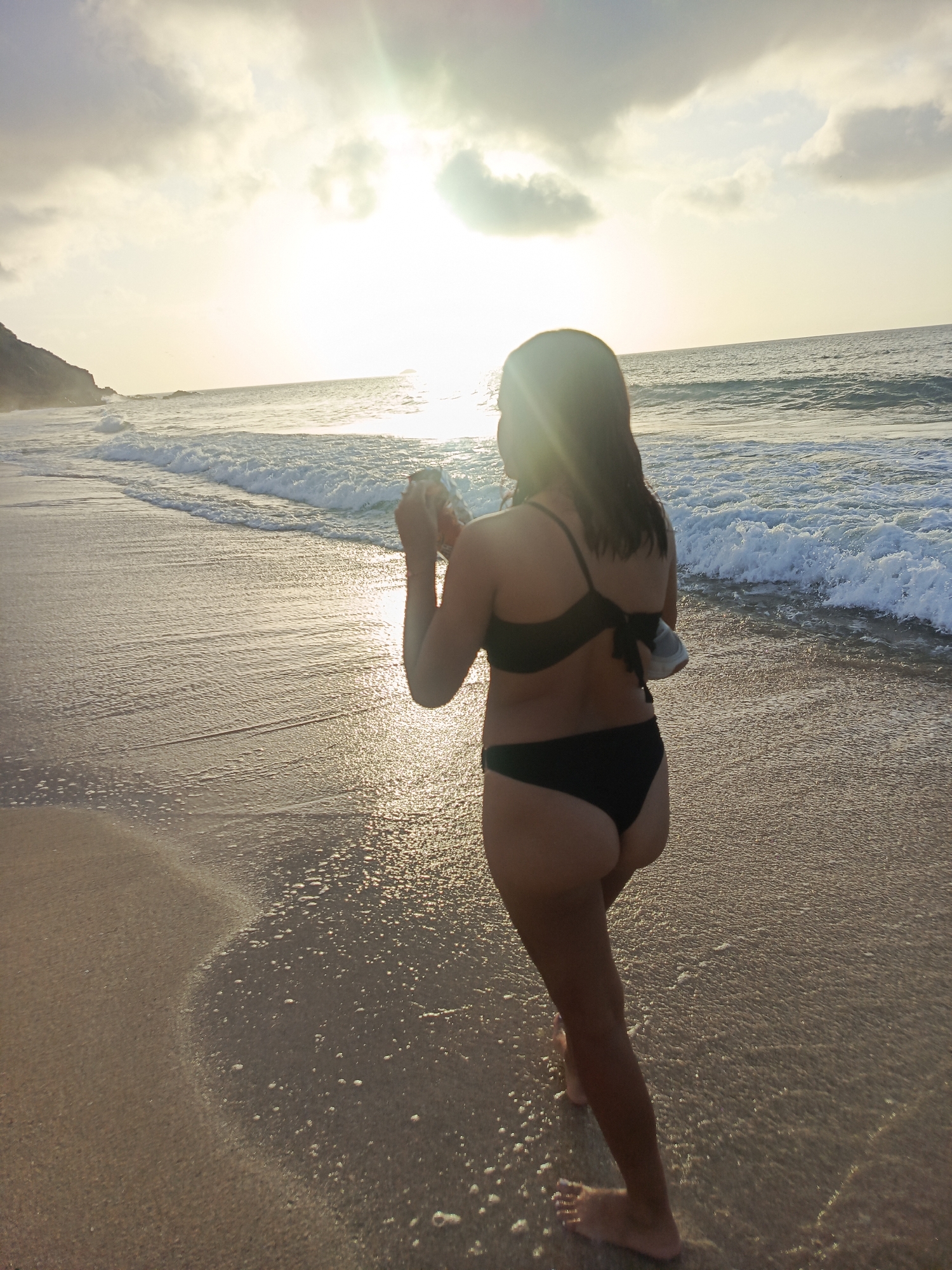 A woman in a black bikini walks along the shoreline of a beach in Pedro González, Venezuela. The sun is setting, casting a golden glow on the water and sand. The waves are gently lapping at the shore, and there are clouds in the sky. The woman is holding a white object in her right hand, and a gray object is tucked into the back of her bikini bottom. Her hair is dark and wet, and she is walking away from the camera.