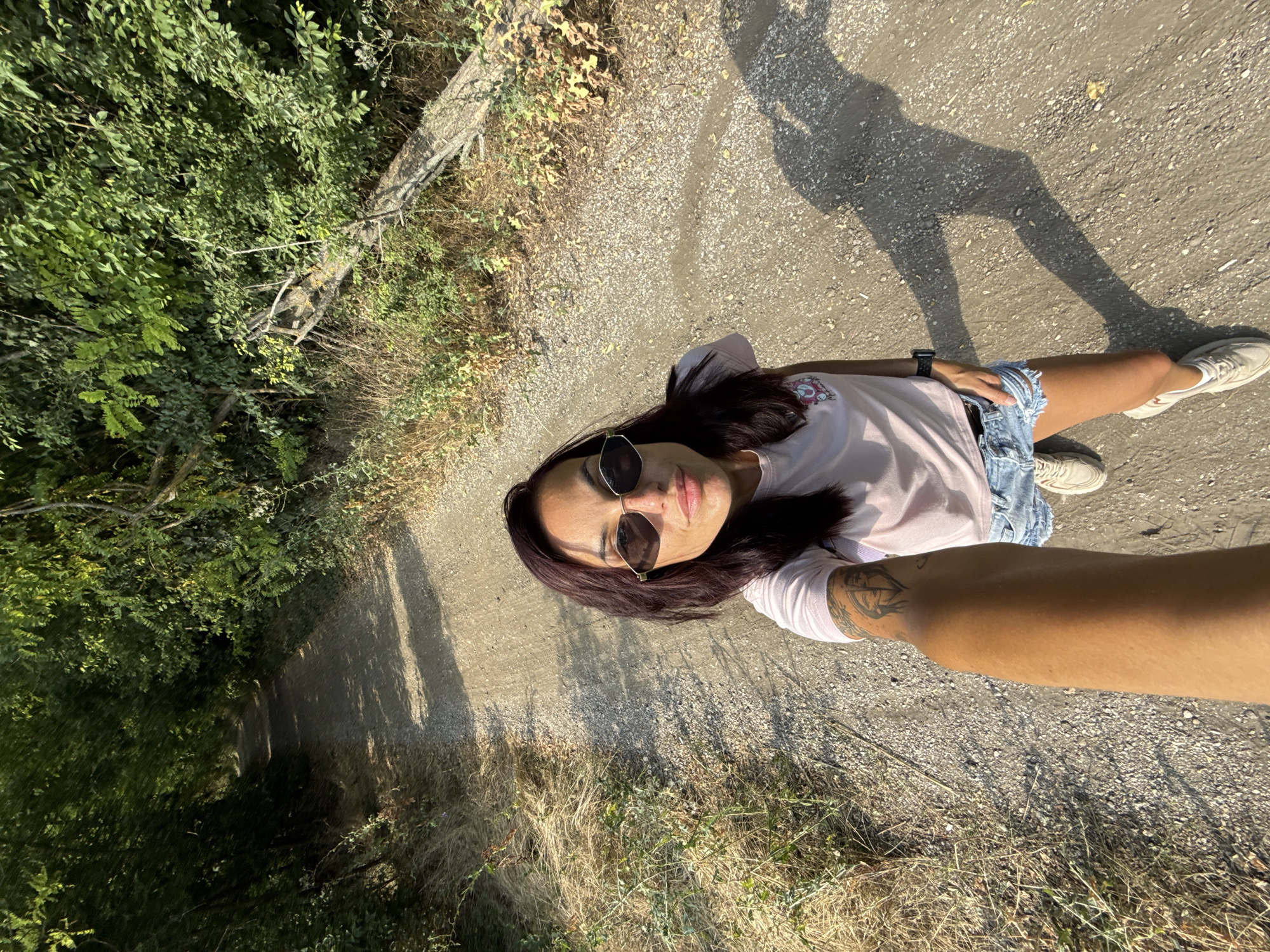 A woman stands on a gravel path in a wooded area. She is wearing sunglasses, a light pink t-shirt, and distressed denim shorts. She has tattoos on her arm and is taking a selfie. The path is lined with trees and dry grass. A fallen tree branch is visible on the right side of the path. The image was captured on a sunny day. The specific location in Pleven, Bulgaria, is not identifiable from the visual information.