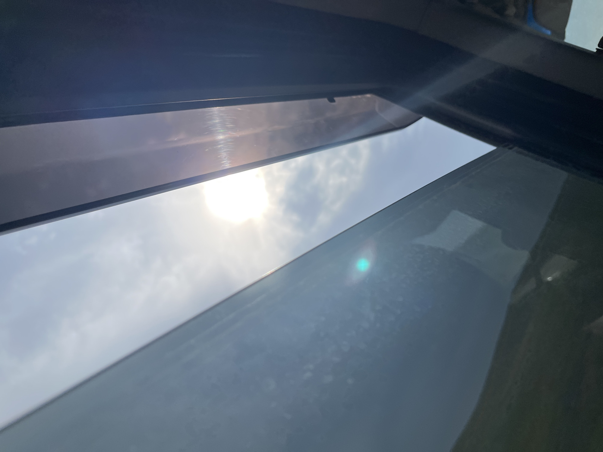 The image, taken from inside a vehicle looking upwards, shows the sun shining brightly through the sky with scattered clouds. The view is framed by the car's sunroof and window. The sun is a dominant feature, causing lens flare that creates a bright white glow and a noticeable green and pink chromatic aberration. Part of the car's interior, likely the A-pillar and roof lining, is visible on the right side of the frame, appearing dark. The left side of the image shows the tinted side window of the car, reflecting some of the outdoor environment, including a hazy green or brown landscape at the bottom. The overall impression is one of bright daylight, likely taken during a car journey.