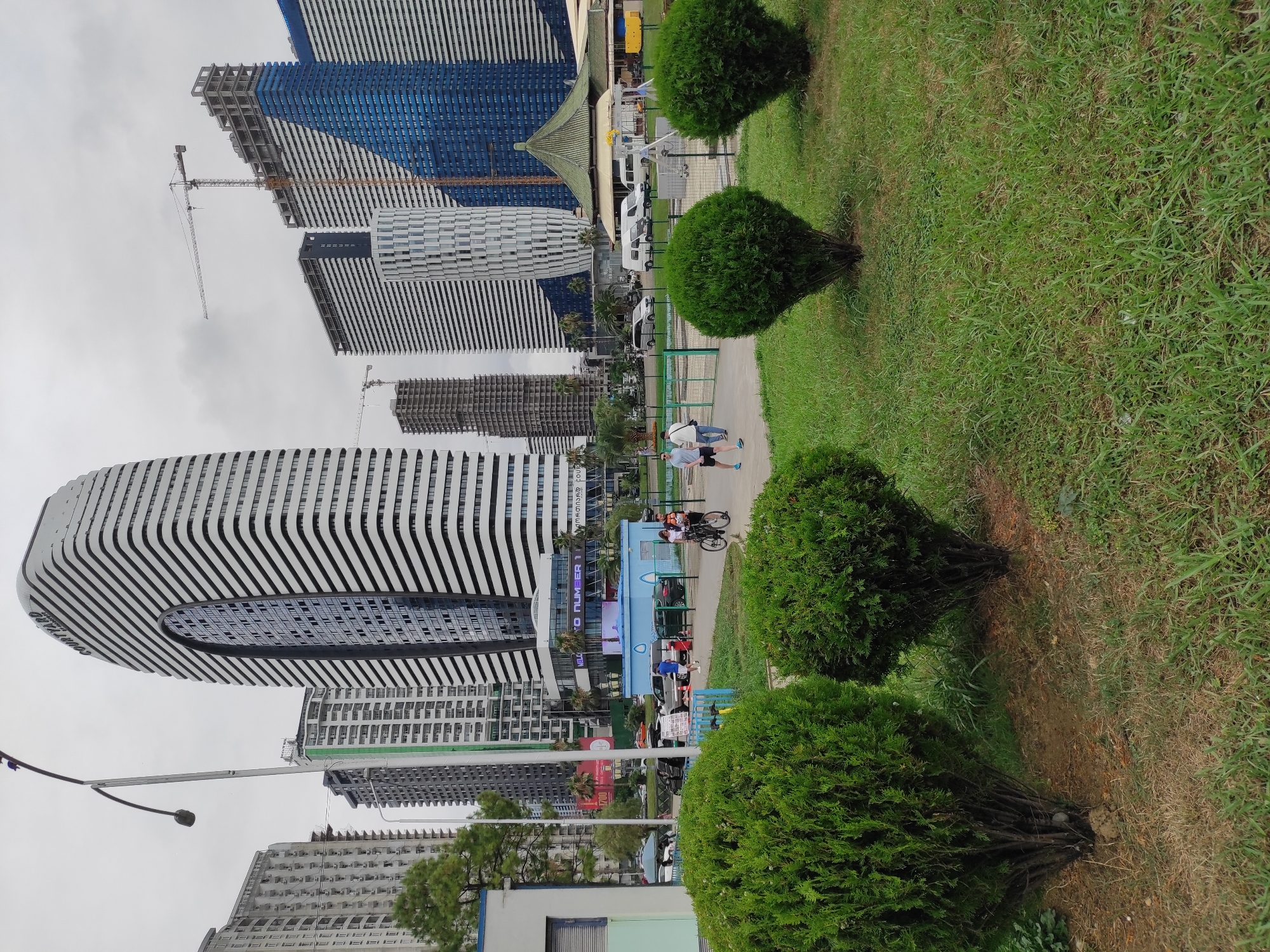 The image depicts an urban street scene with a focus on modern architecture and public space in Batumi, Georgia. In the foreground, manicured green bushes and a grassy lawn create a park-like setting. Beyond the greenery, a sidewalk and a street are visible, with several people walking and a bicycle present. Various vehicles, including cars and vans, are parked or driving on the street.
Dominating the background are several contemporary buildings, characterized by their height and distinctive architectural styles. Notably, a tall, curved building with alternating white and black horizontal bands stands prominently. Adjacent to it are other high-rise structures, some appearing to be under construction with visible cranes, and others finished with facades of white, blue, and glass. A traditional-looking green-roofed building is also visible amongst the modern architecture. The sky is overcast, suggesting a cloudy day. The scene appears to be a bustling urban environment, possibly a commercial or residential area in Batumi, Georgia.