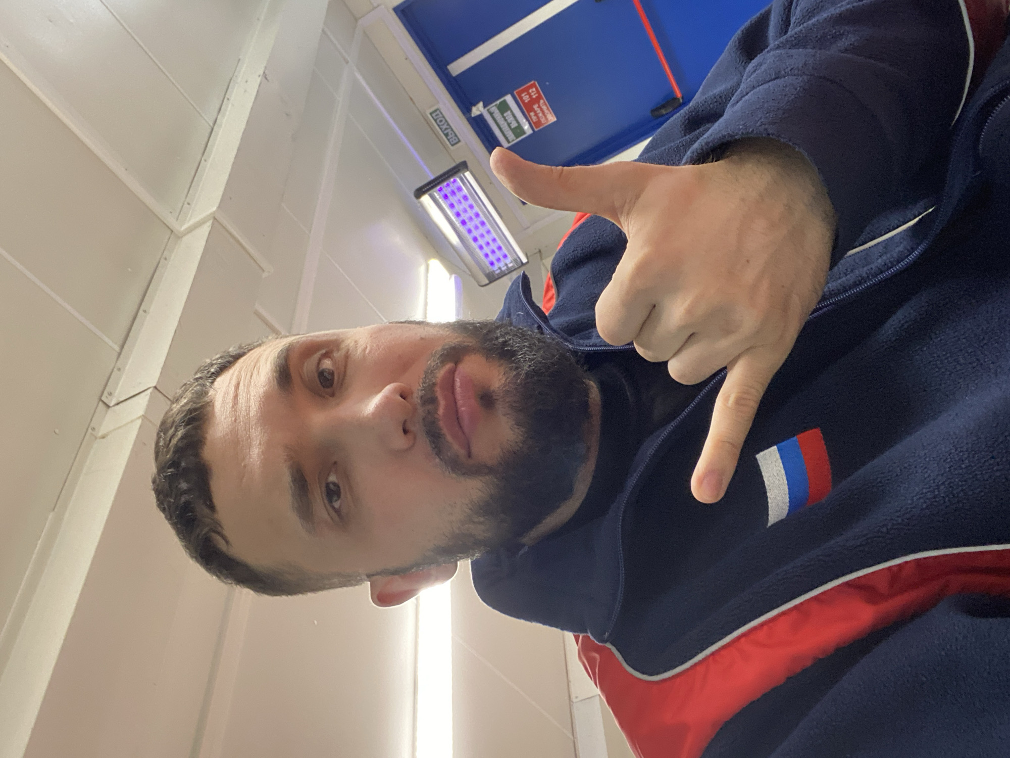 A person with a beard, wearing a navy blue jacket with red trim and a Russian flag patch on the left side, is posing for a selfie. They are making a hand gesture with their thumb up and pinky finger extended. The setting appears to be indoors, under fluorescent lighting, with white and blue walls visible. The location is not mentioned within the image.