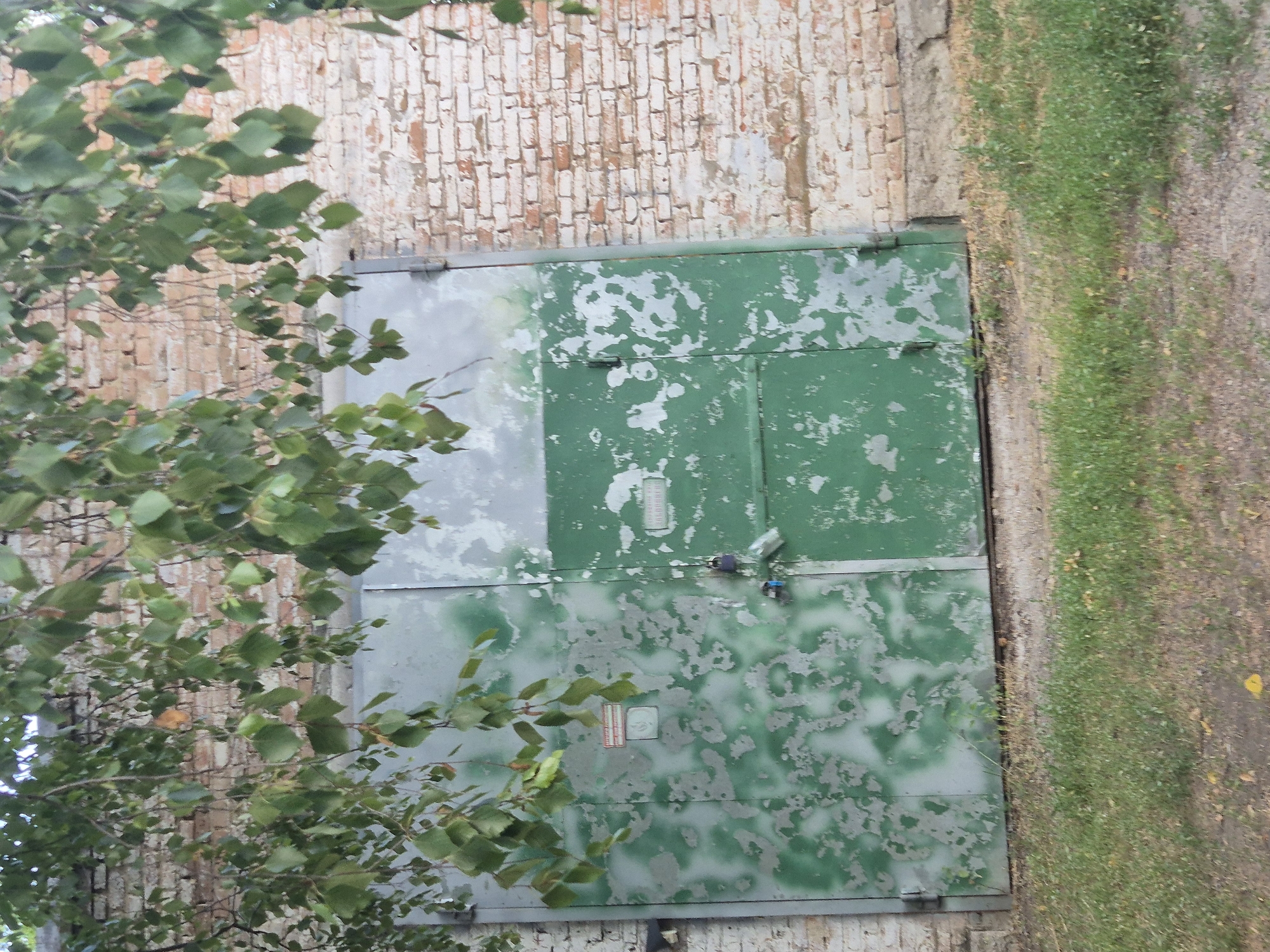 The image shows a weathered, green metal garage door set against a light-colored brick wall. The door is divided into two panels, with the left panel exhibiting significant paint peeling and discoloration, revealing a lighter gray undercoat. The right panel is a more uniform green. Two padlocks are visible on the right door panel, one blue and one silver. A small, faded sign with red and white lettering is affixed to the left door panel. Overhead, branches with green leaves hang down, partially obscuring the top of the garage and the brick wall. The ground in front of the garage is covered with grass and dirt. The overall scene suggests an outdoor setting, possibly in a more rural or industrial area. The location is identified as Sozonivka, Ukraine.