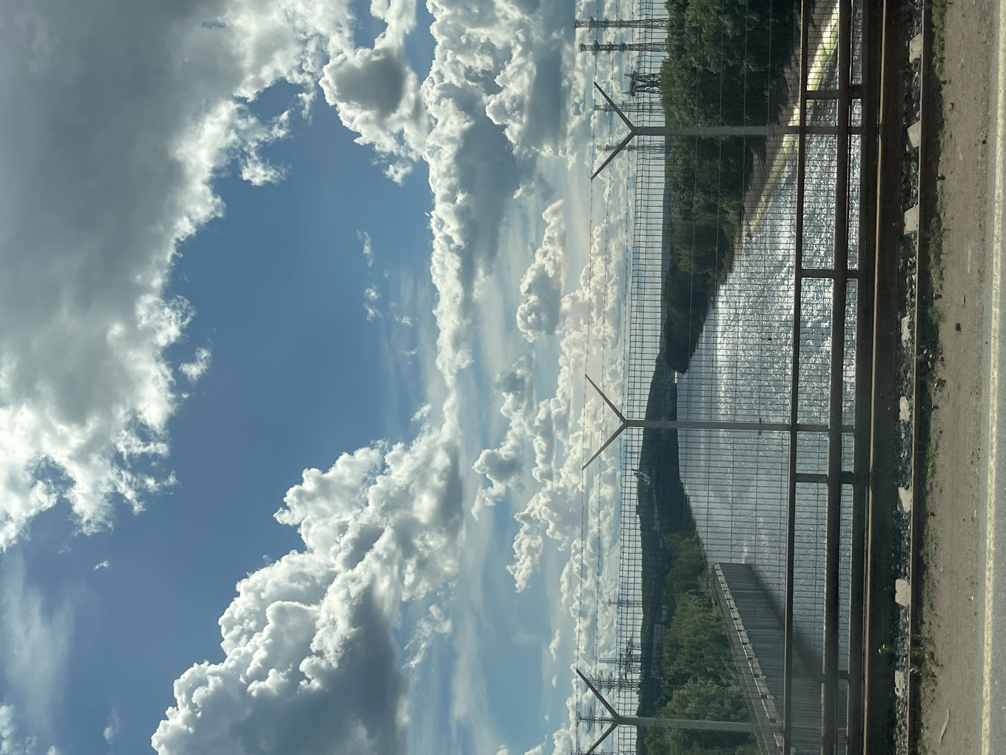 The image shows a partly cloudy sky over a body of water, likely the Kama River, in Chaikovsky, Russia. A metal fence runs parallel to the river. Trees line the opposite bank of the river. Power lines are visible in the distance. The image appears to be taken from a road or walkway.