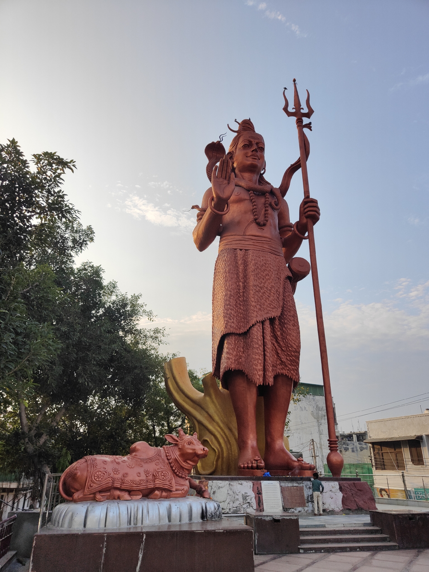 The image depicts a colossal statue of Lord Shiva in Gurgaon, India, with a smaller statue of Nandi, the bull mount of Shiva, in the foreground. The Shiva statue is a reddish-brown color and stands tall against a clear sky with scattered clouds. Lord Shiva is shown with multiple arms, holding a trident in his right hand and raising his left hand in a blessing gesture. A serpent is coiled around his neck, and he wears a beaded necklace and a dhoti-like garment. The Nandi statue is also reddish-brown and is intricately decorated with ornamental patterns. It is positioned on a silver base. To the left of the statues, lush green trees are visible. In the background, there are buildings, some appearing to be under construction or renovation, and a small group of people. The scene appears to be in an outdoor public space, possibly a temple complex or a park.