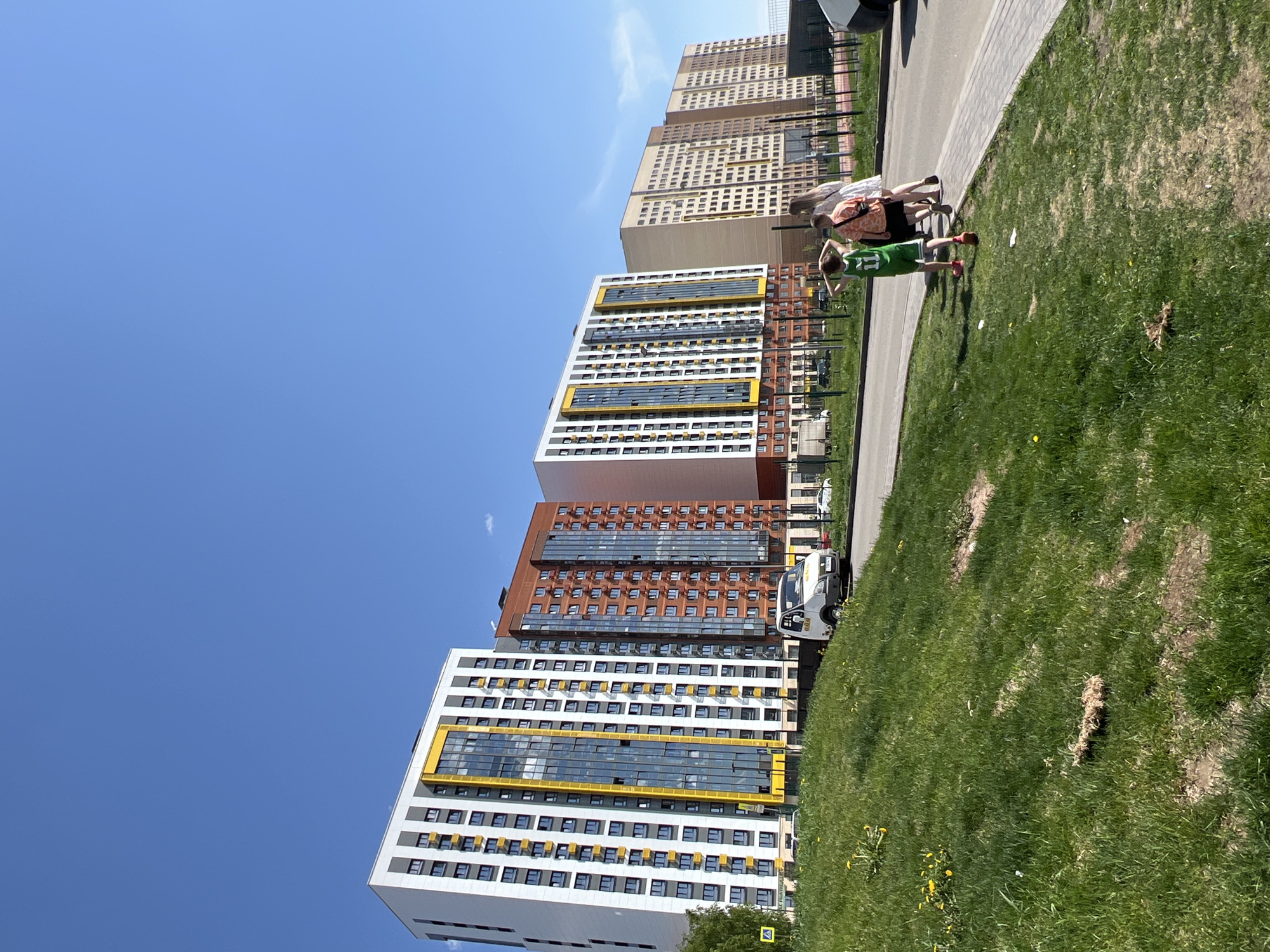 The image shows a sunny day in what appears to be a newer residential area in Izhevsk, Russia. Several modern high-rise apartment buildings with distinct architectural styles are visible. The buildings feature a combination of white, brown, and yellow accents.
Two people are standing on a grassy area alongside a paved walkway. One appears to be an adult woman wearing a printed top and dark shorts. A child, wearing a green jersey with the number 11 and red sneakers, is standing next to her. They seem to be looking at something in the distance.
A white vehicle is parked along the street near the buildings. The sky is a clear blue, suggesting good weather conditions. The setting is urban, with modern architecture and landscaping.