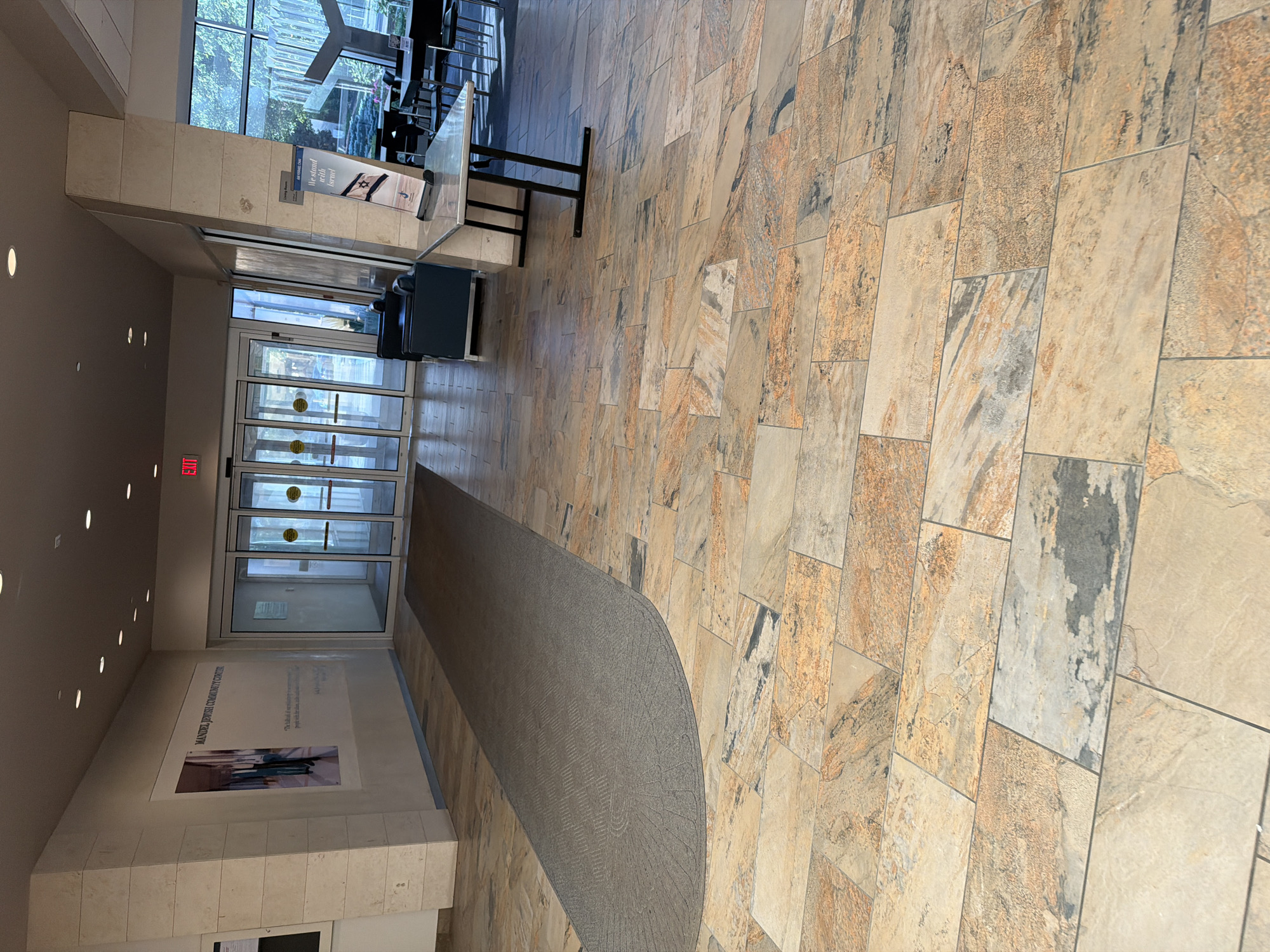 The image shows the interior of the Mandel Jewish Community Center in Beachwood, Ohio, United States of America. The foreground features a floor tiled with large, irregularly shaped tiles in shades of beige, orange, and gray, resembling natural stone. A long, dark gray carpet runner with a subtle patterned texture extends from the left side of the image towards automatic glass doors at the end of a hallway.
To the left, a wall displays a large framed poster or artwork with the text "MANDEL JEWISH COMMUNITY CENTER" and an accompanying image. The wall itself is clad in light-colored, rectangular stone blocks, stacked horizontally.
In the background, the automatic glass doors lead to an outdoor area visible through the glass, with trees and greenery. To the right of the doors, a small seating area includes two dark blue armchairs. Further to the right, a rectangular table with a light-colored stone top is supported by black metal legs. A stand is placed on the table with a banner that reads "We stand with Israel." Beyond this table, in the background, is a seating area with multiple tables and chairs, suggesting a dining or lounge space, with large windows overlooking an outdoor landscaped area with a modern metal sculpture. An "EXIT" sign in red is visible above the automatic doors. The lighting appears to be a combination of recessed ceiling lights and natural light from the windows. There are no people clearly visible in the frame.