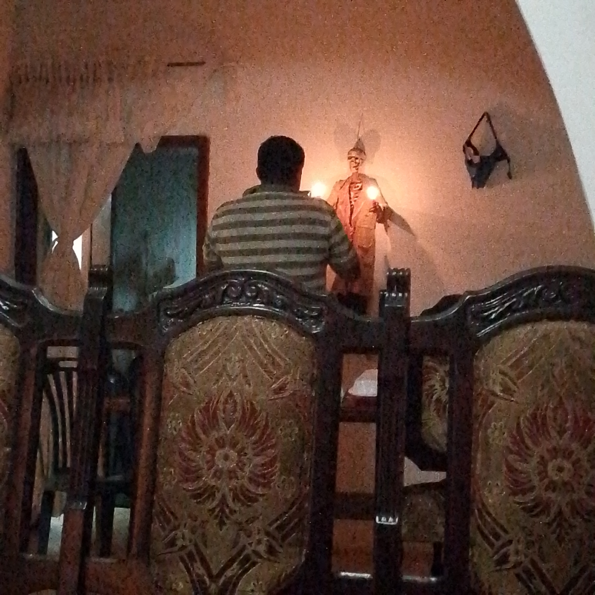 A person in a striped shirt sits in front of a skeleton figure holding lights. The skeleton is wearing a coat and hat. Behind the person is an open doorway with a curtain. A black object, possibly lingerie, hangs on the wall. The scene is dimly lit. The photograph was taken in Tinaquillo, Venezuela. The photograph is taken from behind a chair, obscuring the view.