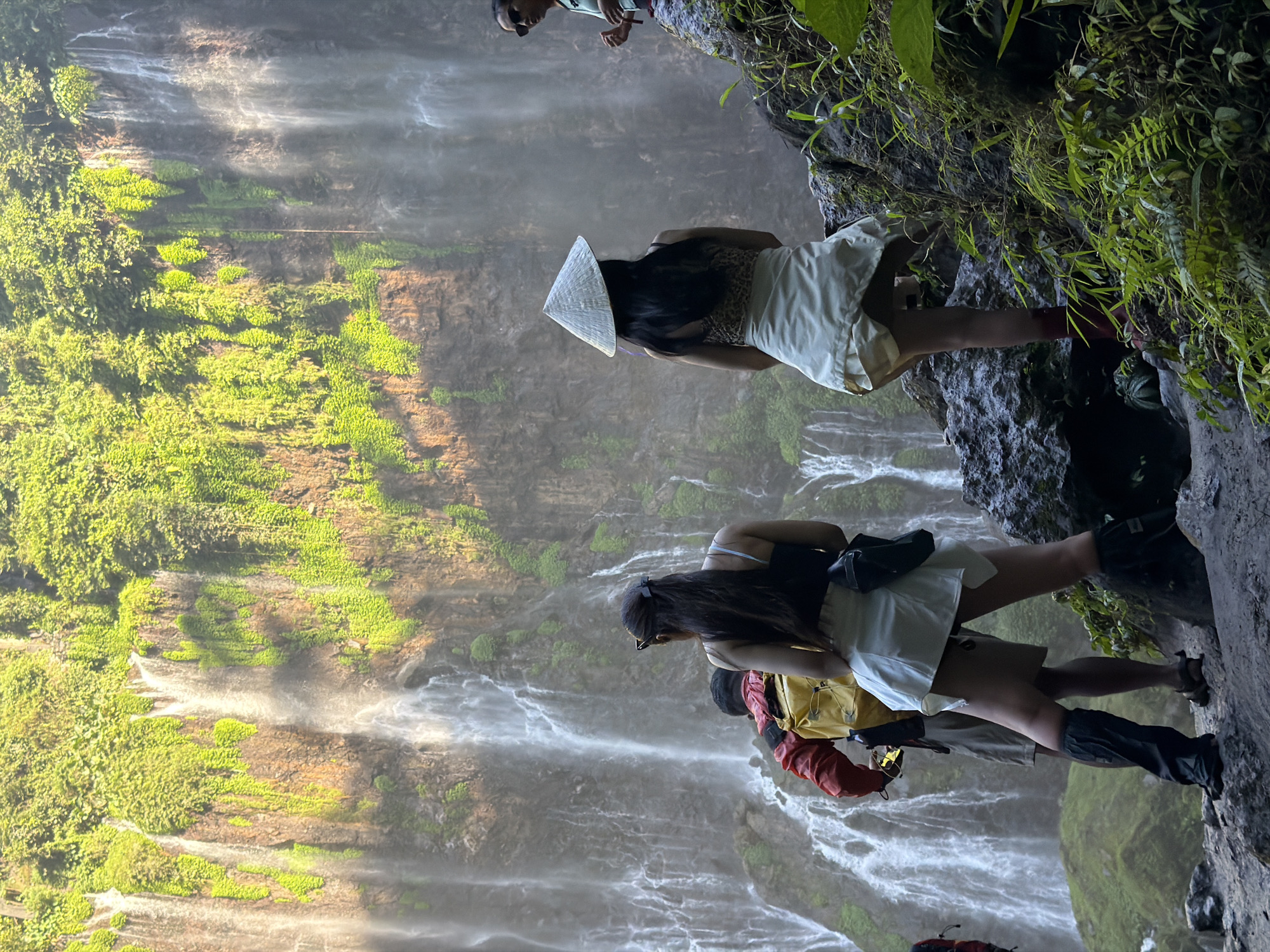 The image depicts a scenic view of a waterfall in Sidomulyo, Indonesia. In the foreground, three individuals are positioned in front of the cascading water. The person closest to the viewer, on the right, is a woman wearing a conical hat and a light-colored skirt, facing away from the camera. To her left, a person in a red jacket and yellow backpack is also facing away. Further to the left, another individual is partially visible. A fourth person, a woman wearing sunglasses, is standing on a rock formation to the far right. The environment is characterized by lush green vegetation covering the rock face behind the waterfall, with numerous streams of water flowing down. The foreground consists of rocky terrain with some green foliage.