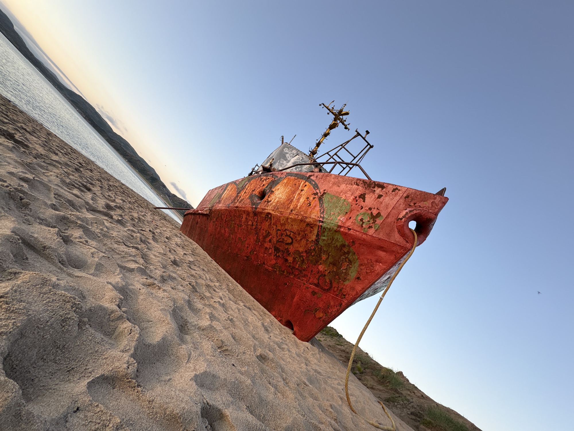An abandoned red ship is beached on the sandy shore of the Barents Sea in Teriberka, Russia. The ship, heavily rusted and partially covered in green algae, lists to its port side. A thick rope is attached to the ship and stretches across the sand. The sky is a clear, pale blue, with a faint haze near the horizon where the sea meets the distant, low-lying mountains. The sand is textured with visible footprints and wave-like patterns. The low angle of the sun suggests it is either early morning or late afternoon, casting soft light on the scene. The overall atmosphere is one of decay and isolation.