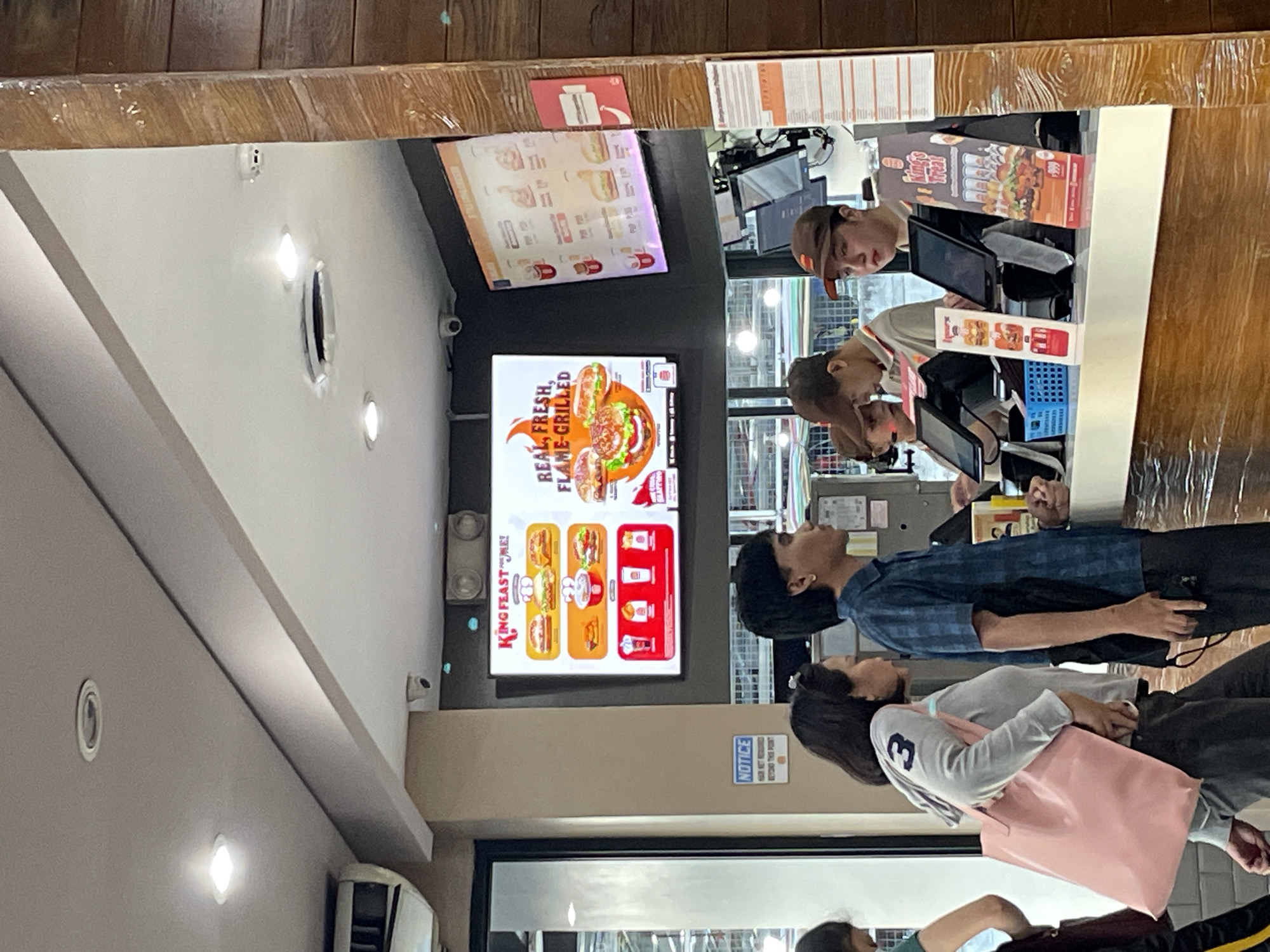 A group of people are at a fast-food counter, likely a Burger King given the signage, in Davao City, Philippines. The counter is equipped with digital menu boards displaying food items and prices, and multiple point-of-sale terminals with touchscreens. Two employees, wearing brown caps and uniform shirts, are visible behind the counter, one attending to a customer. Customers, including a woman carrying a pink tote bag and a man in a blue checkered shirt, are waiting in line. The environment appears to be an indoor commercial space with overhead lighting, a ventilation system, and security cameras. A "NOTICE" sign is posted on a pillar. The overall scene depicts a typical fast-food transaction environment.