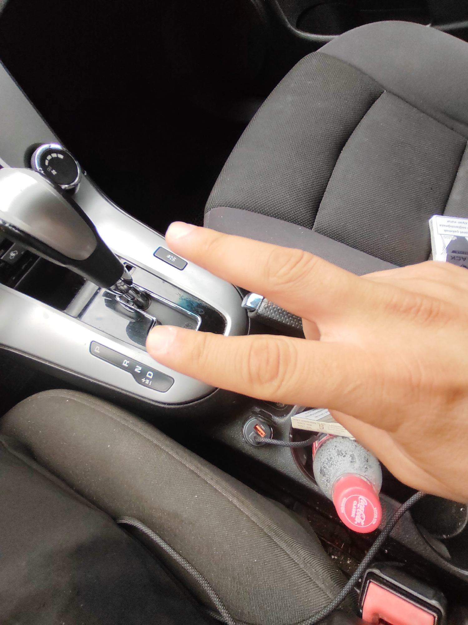 A person's hand is shown making a peace sign gesture in the foreground, positioned over the gear shift of a car. The car interior is visible, including the gear shift mechanism with visible settings for Park (P), Reverse (R), Neutral (N), Drive (D), and a manual shift (+/-). A black fabric car seat is in the background, and a portion of another seat is in the lower part of the frame. A Coca-Cola bottle with a red cap and a cigarette pack are also visible within the car's console area. The scene appears to be inside a vehicle, potentially in Baku, Azerbaijan, given the context of the request.