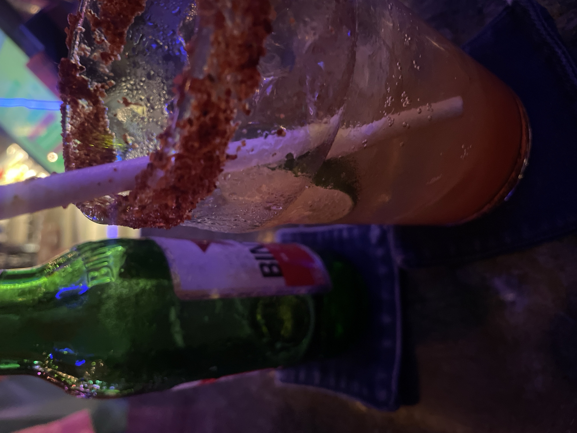 The image, captured in Canggu, Indonesia, presents a close-up, high-angle view of two beverages in a dimly lit, vibrant setting, likely a bar or restaurant during the evening or night.
The primary subject is a cocktail in a clear glass, heavily rimmed with a reddish-brown spice mixture, possibly Tajín or a similar chili-lime seasoning. The drink itself is a reddish-orange hue, filled with ice cubes, several slices of lime, and numerous small bubbles indicating a carbonated mixer. A white straw is partially submerged in the liquid.
Beside the cocktail, a green glass beer bottle is visible. Its label, featuring a white background with a red stripe, shows the partial text "BIN", almost certainly belonging to a Bintang beer bottle, a popular brand in Indonesia. The bottle's surface reflects various ambient blue and green lights from the surroundings.
The background is dark and blurred, with indistinct shapes and soft, colorful light sources contributing to a lively yet intimate atmosphere. A dark fabric, possibly a coaster or part of a tablecloth, is visible beneath the bottles. The scene depicts a relaxed moment of enjoyment, suggesting a social gathering.