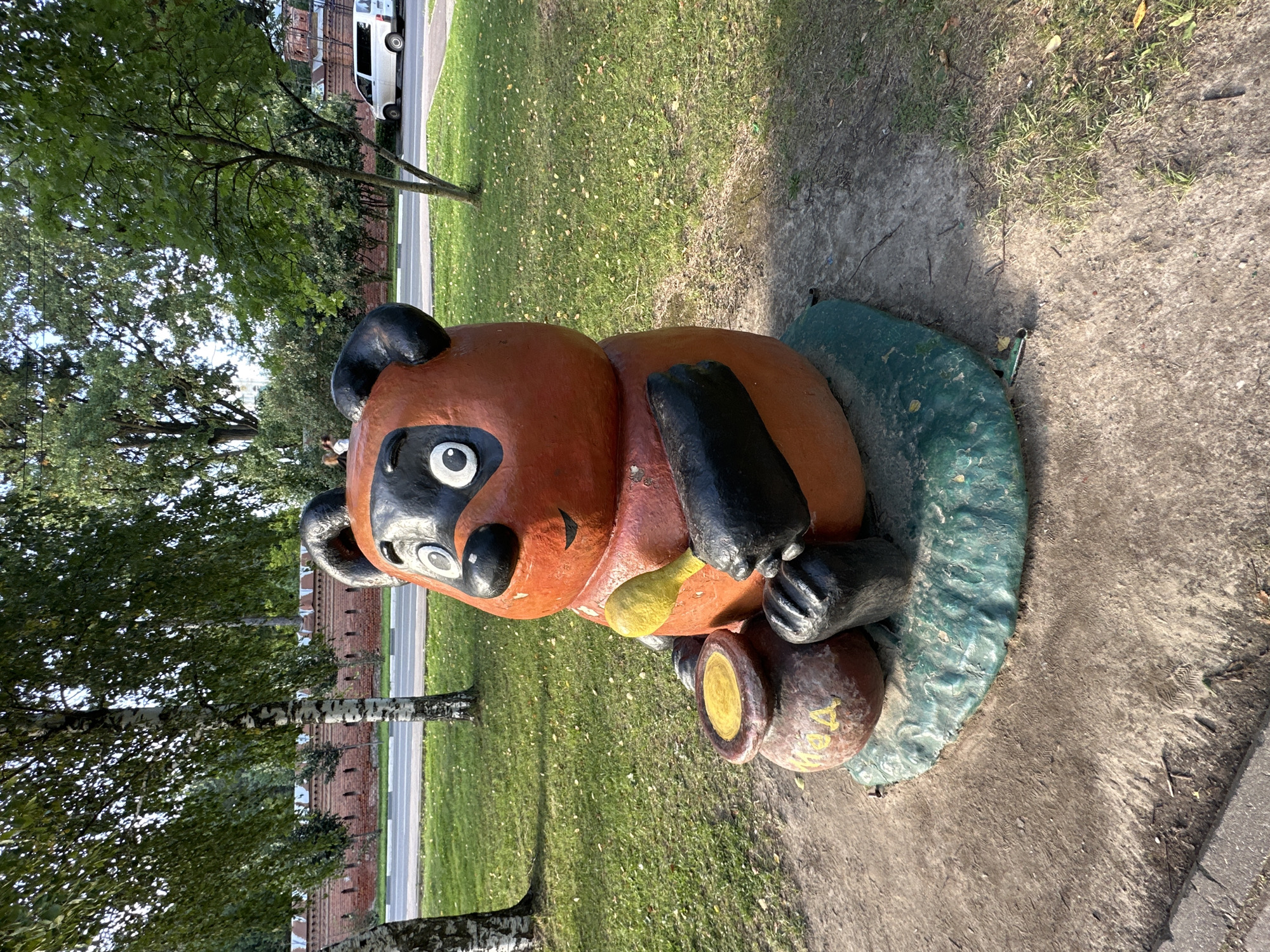A statue of Winnie the Pooh, depicted as an orange bear with black features and big white-rimmed eyes, sits on a green base. He is holding a yellow ladle and a pot of honey, which has the Russian word "МЕД" (honey) inscribed on it. The statue is located in a grassy area, with trees and a brick wall in the background. A white van is parked on a road behind the wall. The statue is situated near a sidewalk and a paved area. The overall setting suggests an outdoor public space, likely a park or recreational area within Sergiyev Posad, Russia, given the presence of Russian text and architectural style.