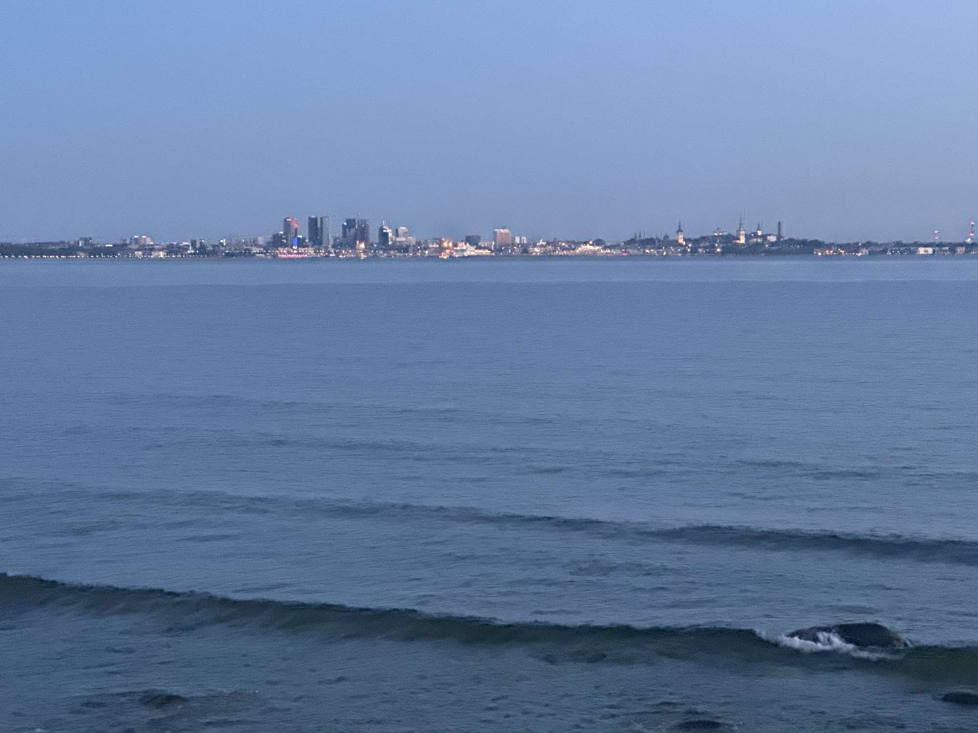The image displays a wide-angle, low-light scene of the city of Tallinn, Estonia, observed across a body of water, likely Tallinn Bay. The sky, occupying the upper third, is a clear, uniform dark blue. The lower two-thirds consist of dark blue-grey water with subtle ripples and several visible waves; a prominent wave breaks towards the bottom right, partially revealing a dark, submerged rock.
The illuminated city skyline stretches horizontally across the horizon. In the left and center-left sections, a cluster of modern high-rise buildings with distinct artificial lighting, some displaying colorful hues, forms a contemporary urban district. To the right of this, the skyline transitions to the historic silhouette of Tallinn's Old Town, characterized by multiple prominent spires and towers, which are also illuminated. Further to the right, the urban sprawl continues with scattered lights and a distinct red-and-white striped structure, possibly a chimney or communication tower. The entire city edge along the water is delineated by numerous small, glowing lights. No individuals or specific vehicles are discernible within the frame.