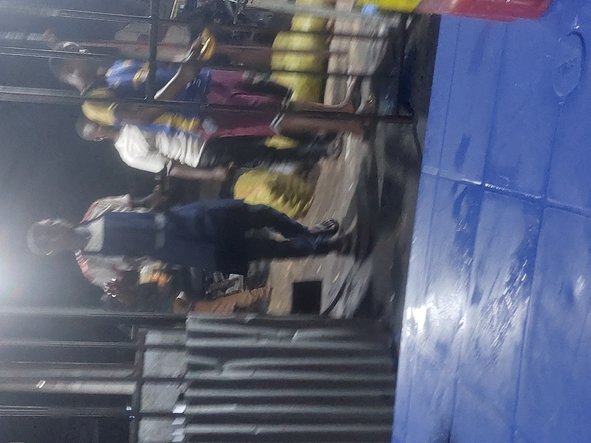 A dimly lit outdoor scene shows a group of people near what appears to be a shop or market stall. Several individuals are visible, some standing and interacting with the items on display. The scene suggests an informal setting. The location is likely Dar es-Salaam, Tanzania, based on the prompt's instructions, but specific details within the image do not confirm or deny this. The environment includes corrugated metal structures and what appear to be plastic items, perhaps for sale or storage.