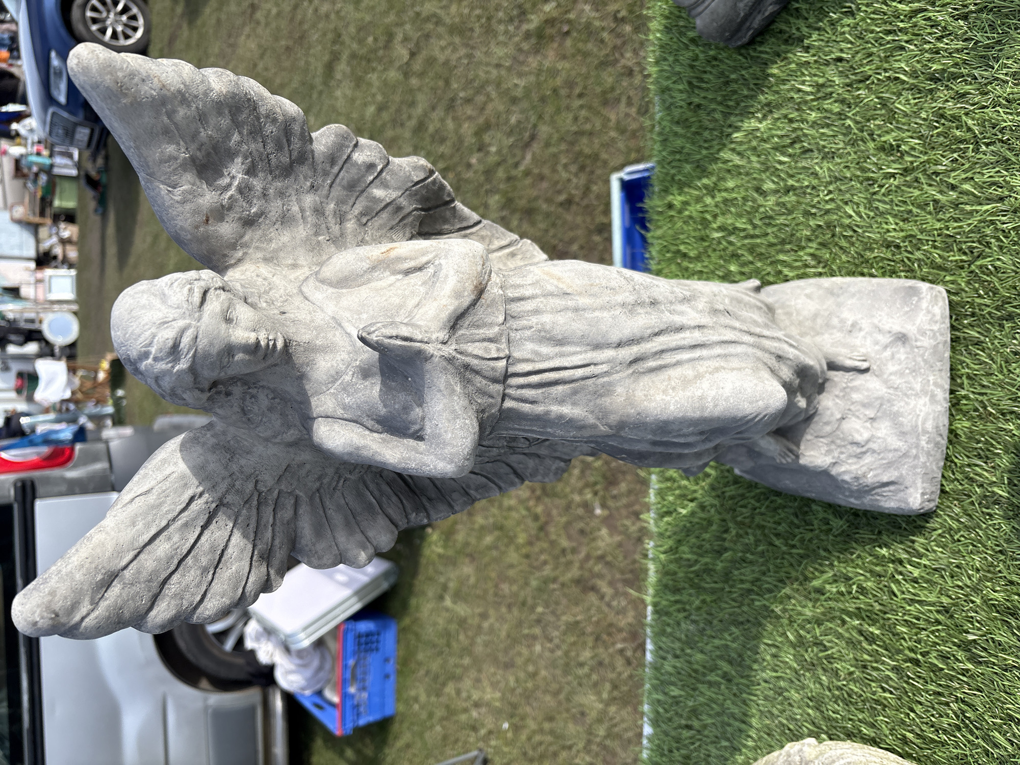 A statue of an angel with large wings stands on a rectangular base. It is made of gray stone and appears weathered. The statue is positioned on a patch of artificial green turf. To the left, out-of-focus, are parts of a vehicle and various items, suggesting a possible market or sale environment. In the background, more grass and another vehicle are visible. The setting is likely an outdoor space in Chirk, United Kingdom.