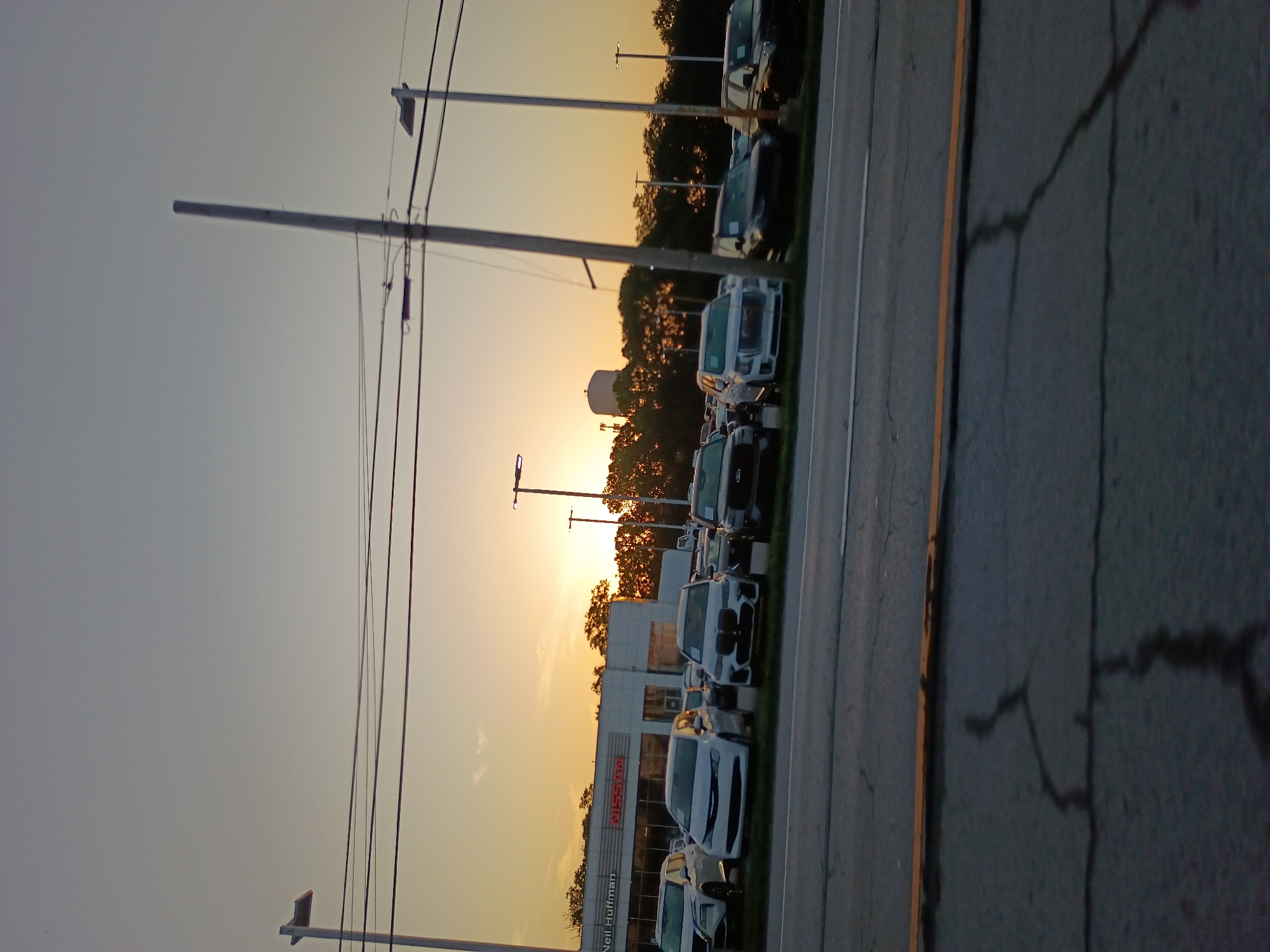 This image captures a car dealership with a row of vehicles parked in front of a building. The sun is setting, casting a warm glow across the sky and illuminating the scene with a golden light. Trees line the background, and a water tower is visible in the distance. The sky is clear, suggesting pleasant weather. A utility pole with wires stands tall in the foreground, adding to the urban setting. The cars appear to be new models, with one prominently displaying the "Nissan" logo. The name "Neil Huffman" is also visible on the building, likely indicating the dealership's name. The overall mood is serene, with the sunset creating a peaceful atmosphere.