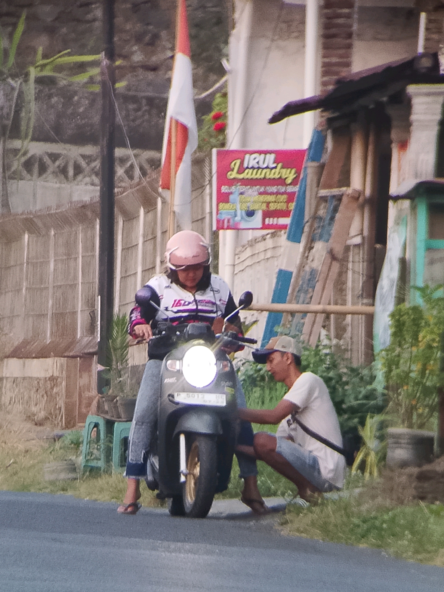 In Jember, Indonesia, on a paved road, a woman wearing a pink helmet and a patterned jacket rides a dark-colored scooter. A man in a white t-shirt and light-colored shorts, wearing a baseball cap, kneels beside the scooter, appearing to adjust something on its front. The scooter has a license plate with the characters "P 5013 HG". In the background, a red and white Indonesian flag is visible on a pole. A pink sign advertising "IRUL Laundry" is also present. The environment suggests an outdoor setting with some vegetation and buildings.