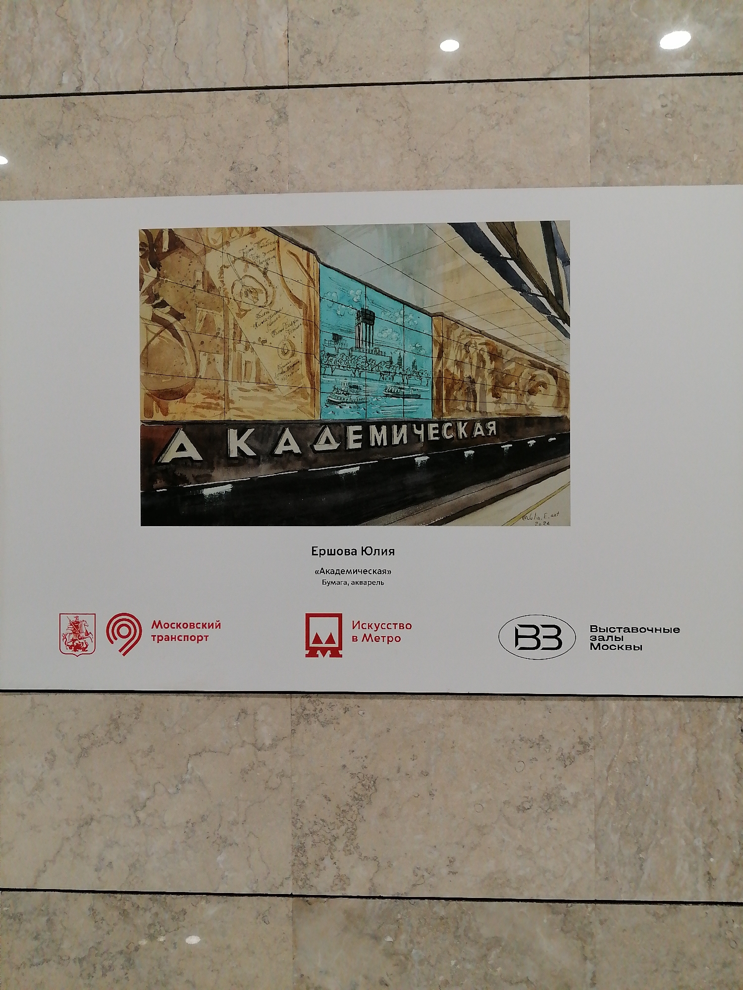 The image is a photograph of a sign featuring a watercolor painting of the "Akademicheskaya" metro station in Moscow, Russia. The sign is mounted on a wall with a light beige marble pattern.
The watercolor painting depicts a segment of the metro platform with the station name "Akademicheskaya" prominently displayed in Cyrillic letters. The station walls are decorated with murals in shades of brown and blue-green. On the left, the murals feature historical maps and documents, while the blue-green section shows images of the Neskuchny Garden, a historical park located near the station. A signature, "Julia E art 2026," is visible in the lower right corner of the painting.
Below the painting are several logos. On the left is the logo of "Moscow Transport." In the center is the logo of the "Art in Metro" project. On the right is the logo "Exhibition Halls of Moscow."