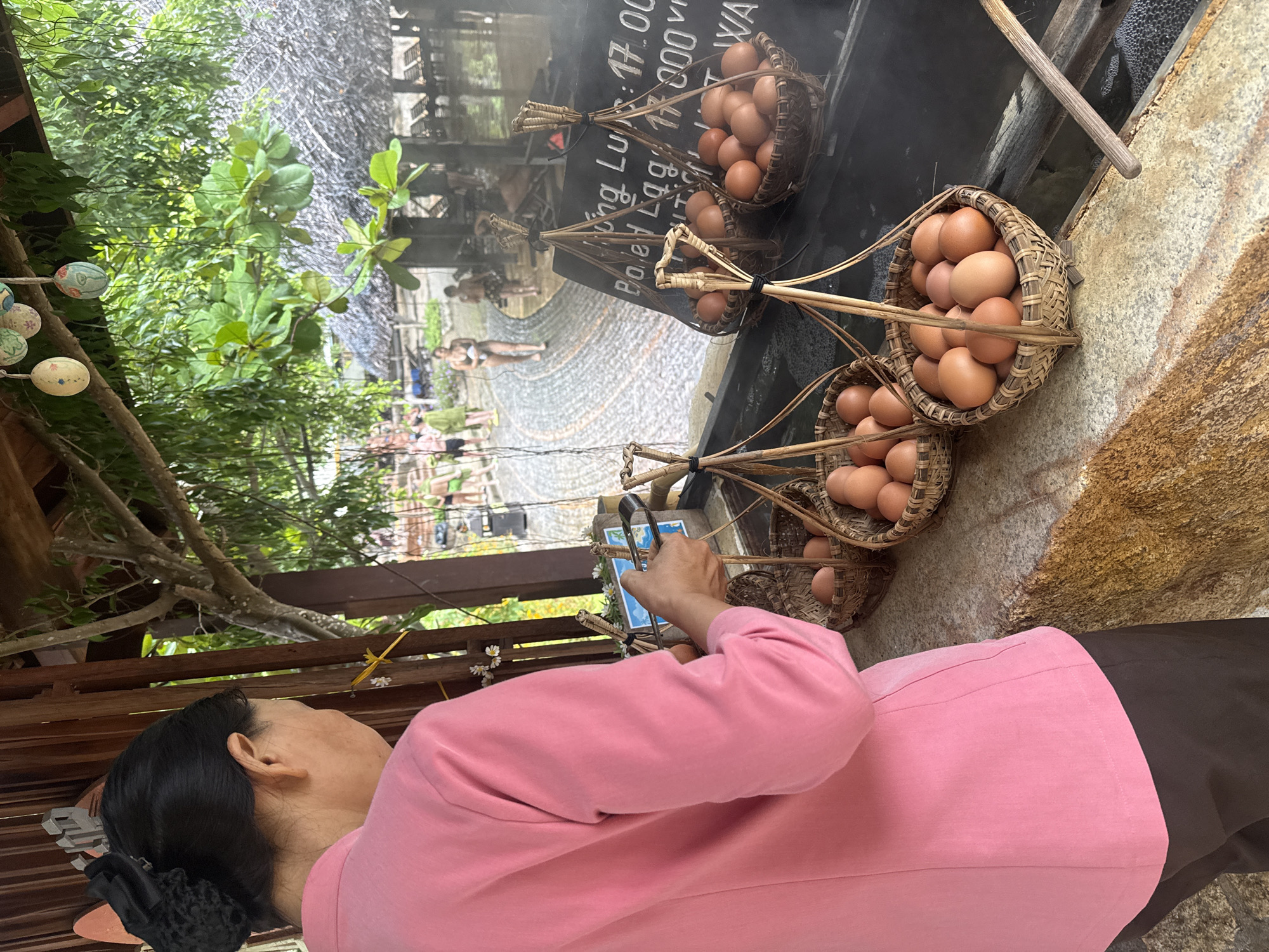 In Nha Trang, Vietnam, a person wearing a pink top and dark pants stands with their back to the viewer, holding a pair of tongs. They appear to be preparing to place or retrieve brown eggs from a woven basket. In front of them, several baskets are filled with similar eggs. Behind the person and to the right, a dark surface with steam rising suggests a hot spring or cooking area. A sign with prices written on it is visible, indicating "Boiled Eggs" and "17,000 VND". In the background, other people are visible near a body of water, and thatched-roof structures are present. The environment is outdoors, with trees and vegetation overhead.