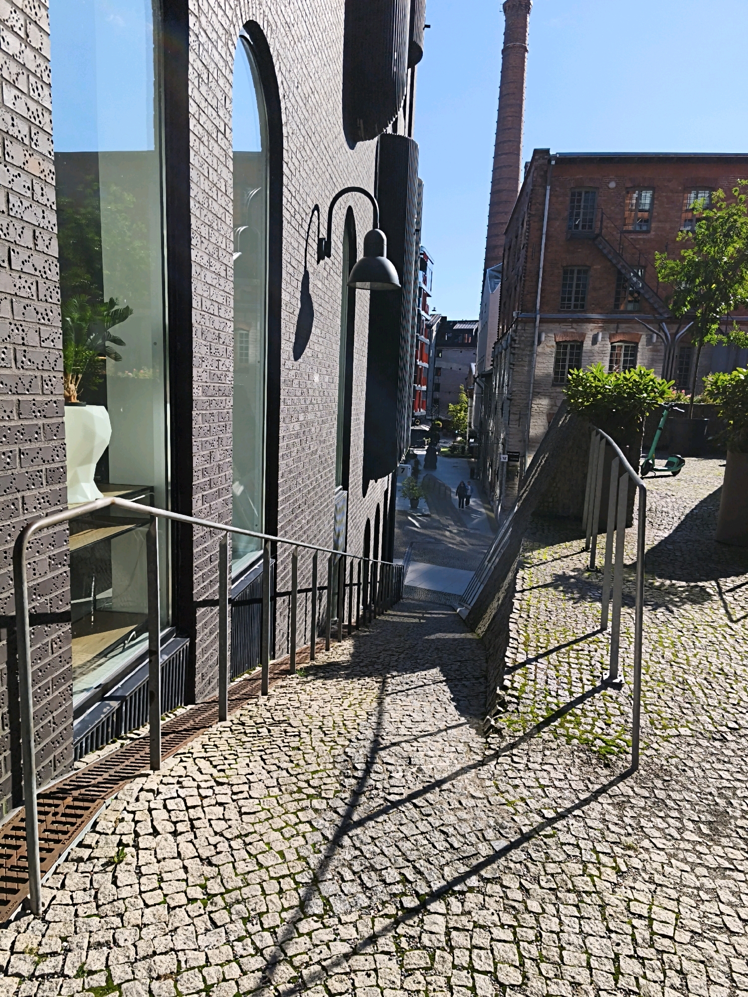 This is a ground-level, eye-level shot of a cobblestone pathway descending between brick buildings in Tallinn, Estonia. The pathway is lined with metal railings on both sides and is partially covered in sunlight and shadows cast by the surrounding structures. On the left, a modern brick building with large windows reflects the blue sky and greenery. On the right, an older brick building with a tall, red brick chimney rises in the background, suggesting a former industrial area. Two figures are walking down the pathway in the distance. A green electric scooter is parked on the right side of the pathway near a tree. The overall scene depicts a street in a historic yet revitalized urban area.