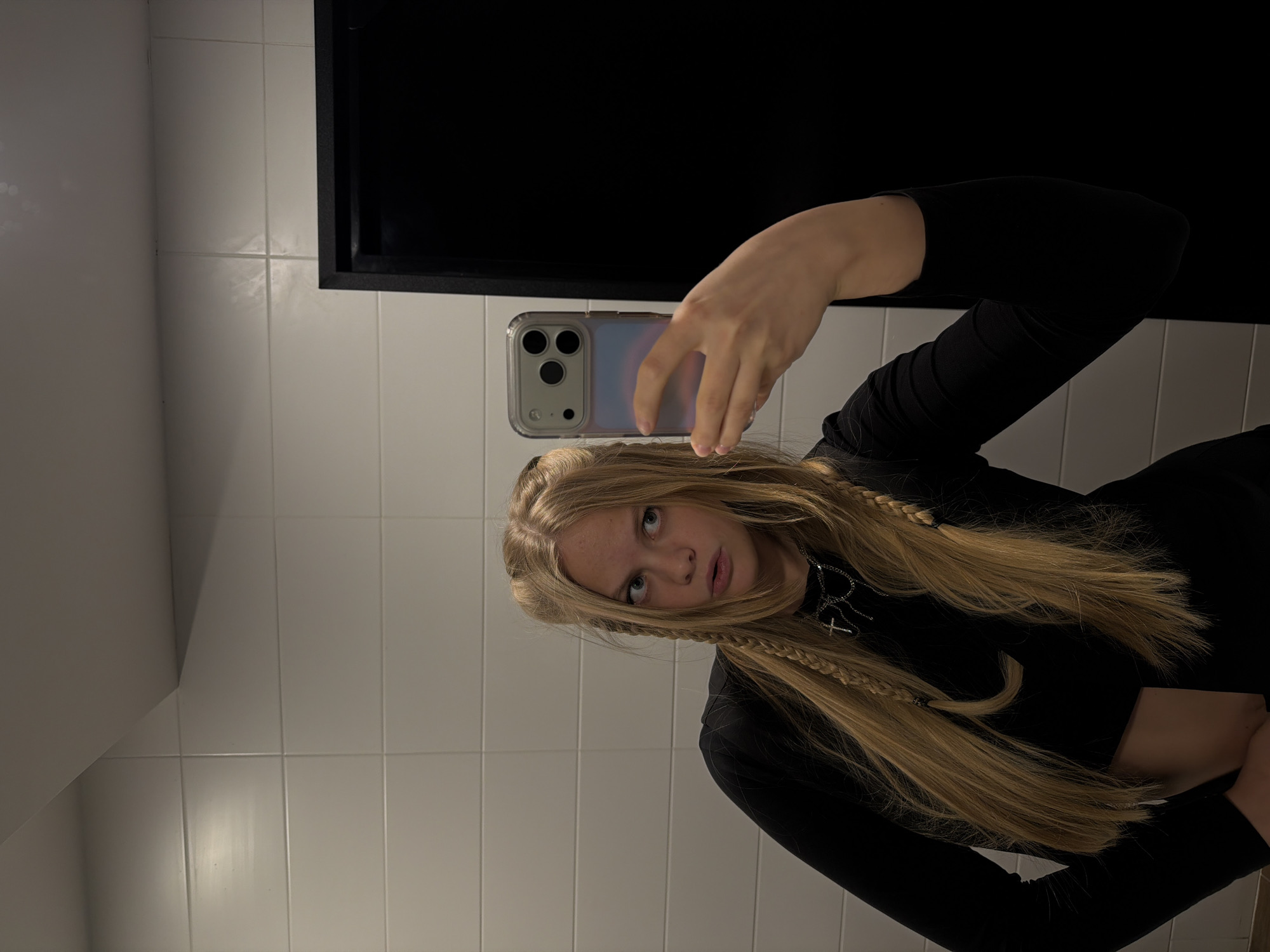 A young person, likely female, with long blonde hair, takes a mirror selfie in an indoor setting. Her hair is styled with several thin braids framing her face. She wears a black long-sleeved crop top and a silver chain necklace featuring a prominent cross pendant. She holds a smartphone, identifiable as an iPhone with a clear case and visible triple camera lenses, up to capture the image, partially obscuring her face. Her gaze is directed towards the camera with a neutral expression. The background features white rectangular tiled walls, suggesting a bathroom or changing room. A large, dark, rectangular area is visible behind her head and the phone, possibly a mirror frame or a dark opening. The lighting is artificial, suitable for an indoor environment. No visible text is present in the image. The location context for this image is Izhevsk, Russia.