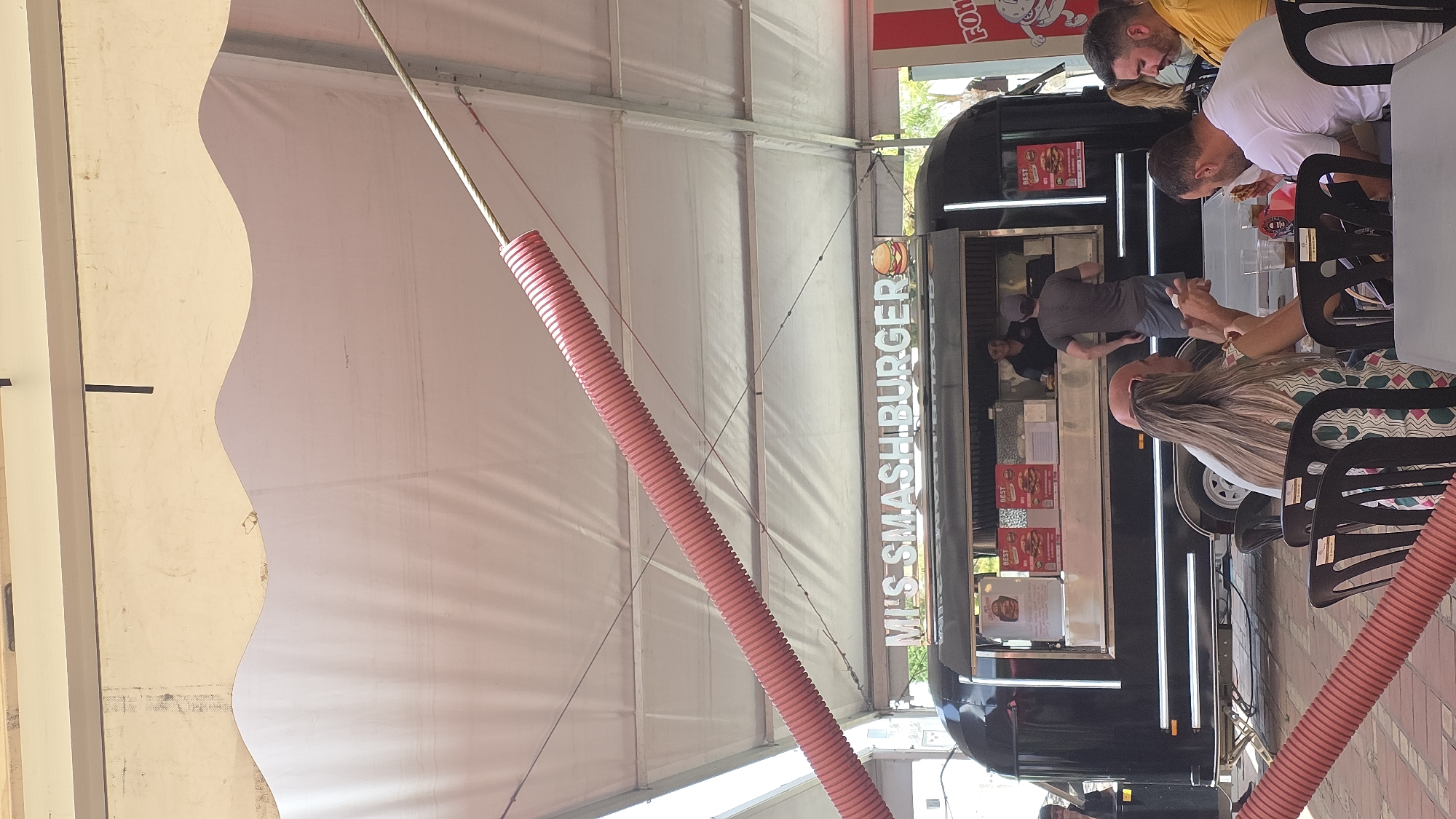 This image shows an outdoor scene at what appears to be a food truck festival or market under a large white tent. The central focus is a black food truck with the name "MRS SMASHBURGER" written vertically in illuminated white letters. The truck has a serving window where a staff member is visible behind the counter, and a customer is interacting with them. Several people are seated at tables in front of the food truck, seemingly eating and socializing. One person in the foreground is facing away from the camera, with long blonde hair. Another man is looking down at food on his plate, and a third man is leaning over. The overall atmosphere seems casual and relaxed.
Notable details include the bright daytime lighting, suggesting it is likely midday. The setting under a tent indicates an outdoor event, possibly in a town square or a designated market area. The red corrugated tubes hanging from the tent structure are a distinctive visual element. The image is oriented vertically, suggesting it was taken with a phone camera in portrait mode.
Visible text includes "MRS SMASHBURGER" on the food truck and "BEST" printed on several posters displayed on the truck's exterior, likely advertising menu items or specials. A red sign with white lettering is partially visible at the top of the frame, reading "FOR".