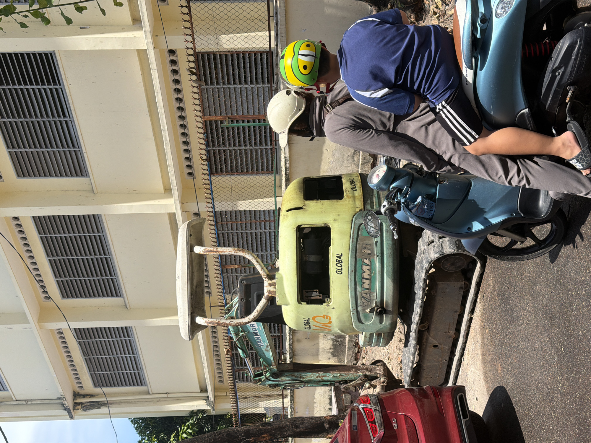 In Nha Trang Ward, Vietnam, an outdoor scene features a light yellow building with louvered windows. In front of the building, a small, green excavator with "YANMAR" and "GLOBAL" branding is parked. To the right of the excavator, two individuals are seated on a light blue scooter. The person in front wears a beige helmet and a dark gray jacket, while the person behind wears a green, yellow, and black helmet and a blue shirt with white accents. A red car is parked to the left of the excavator. The ground is paved with asphalt and dirt.