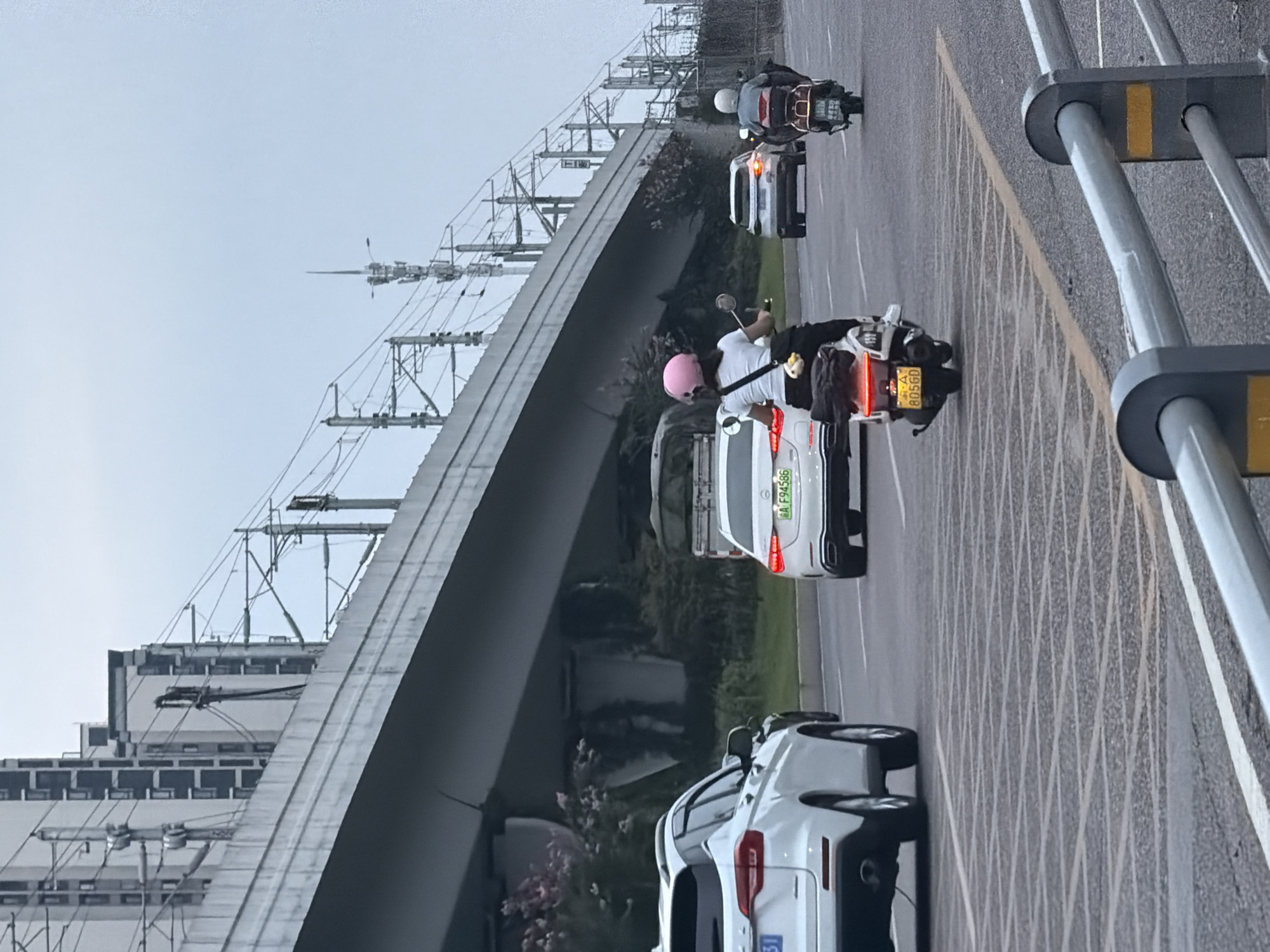 This is a full-shot, eye-level, medium-distance photograph taken on a cloudy day in Jiangjin, China. The scene is a multi-lane road with vehicles driving in the same direction. In the foreground, a white SUV is on the left, and a white sedan is to its right. Further ahead, there are two motorcycles. The rider of the first motorcycle, a woman wearing a pink helmet and a white shirt, is facing away from the camera and appears to be gesturing. The second motorcycle is ahead of her and to the right, with a rider also facing away from the camera. In the background, a large elevated highway structure dominates the scene, with electrical lines and poles visible above it. Buildings, also in the background, are partially obscured by the highway. The road is paved and marked with lane lines, including a distinctive diamond-shaped pattern on the right side of the lane.