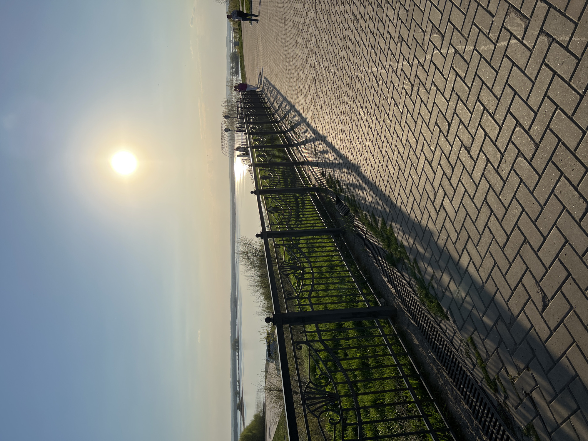 The image captures a horizontal view of a walkway in Sviyazhsk, Russia. The scene is illuminated by the sun, positioned on the left side of the frame, which casts long shadows of the wrought iron fence onto the paved walkway. The walkway is constructed of gray brick in a herringbone pattern.
A grassy area borders the walkway, enclosed by the wrought iron fence. Beyond the fence, a body of water is visible, with distant land visible in the background. The sky is clear and pale blue, merging with the horizon. Two figures are walking in the distance. Another figure leans on the rail.